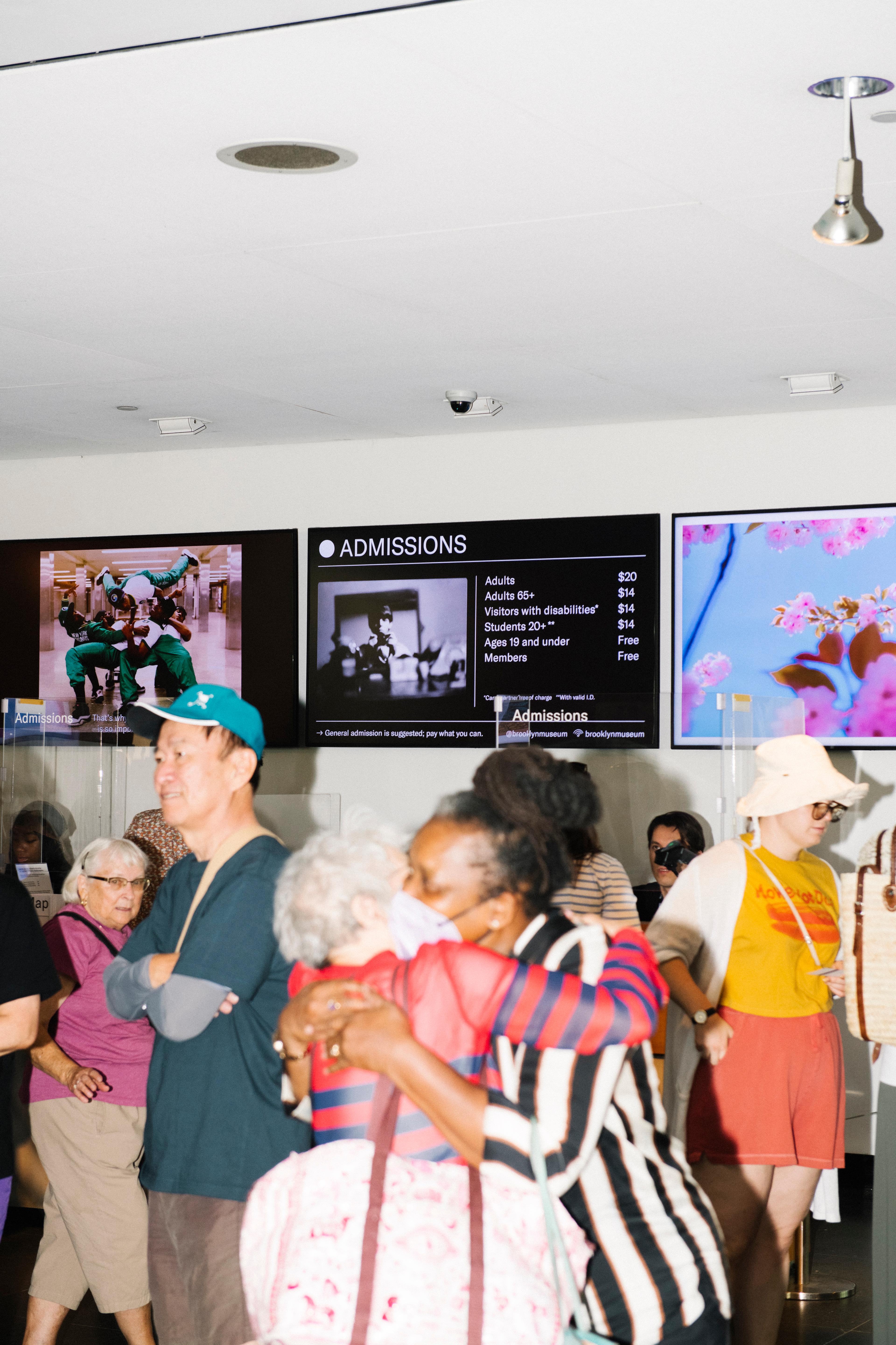 Two older adults wearing masks hug in front an admissions desk, surrounded by other people