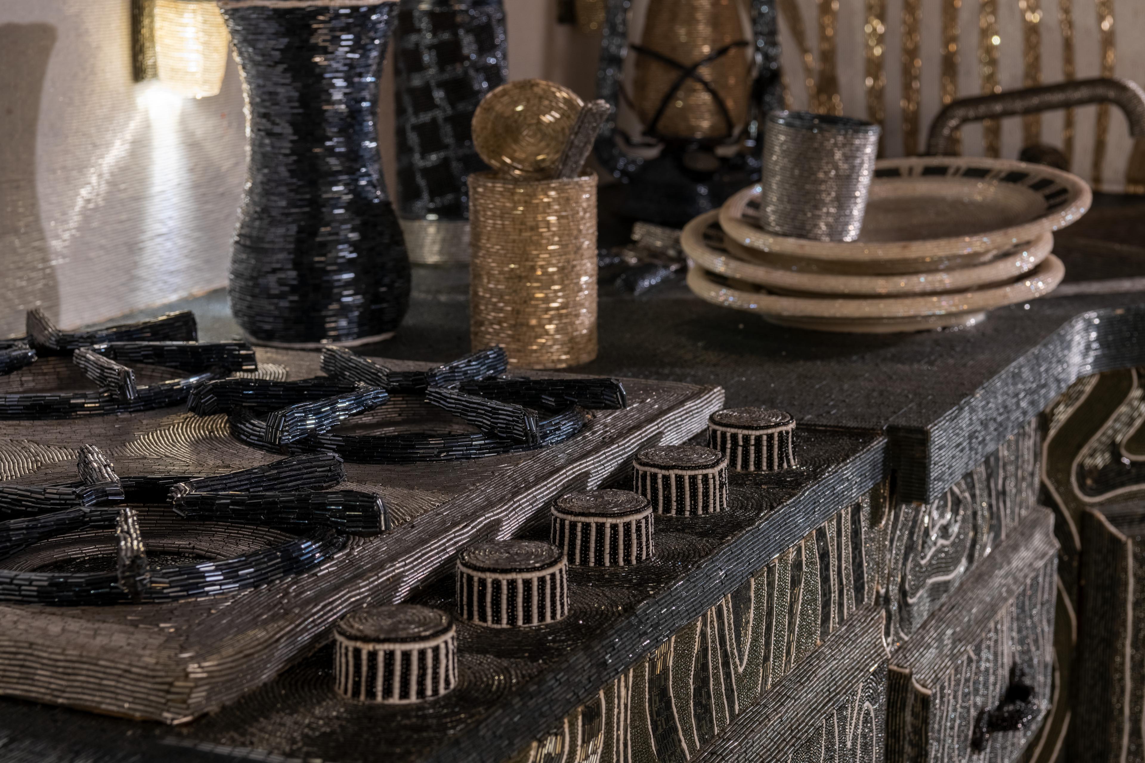 Interior detail of a kitchen scene showing a stove, and a stack of dirty dishes at the sink. Everything is made of beads.