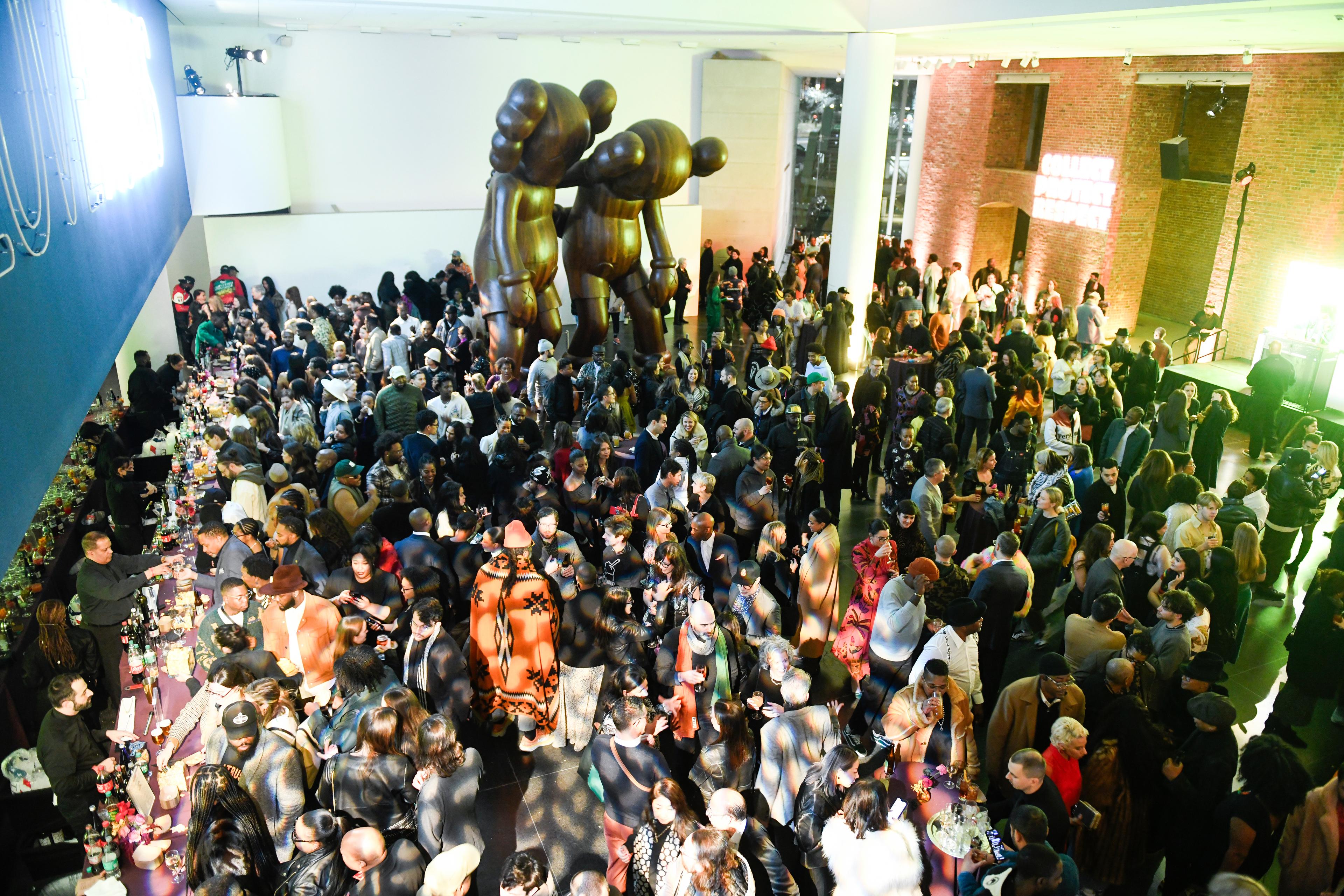 Hundreds of people crowd a large room for an event where there is a bar, a stage, and two large sculptures in the background.