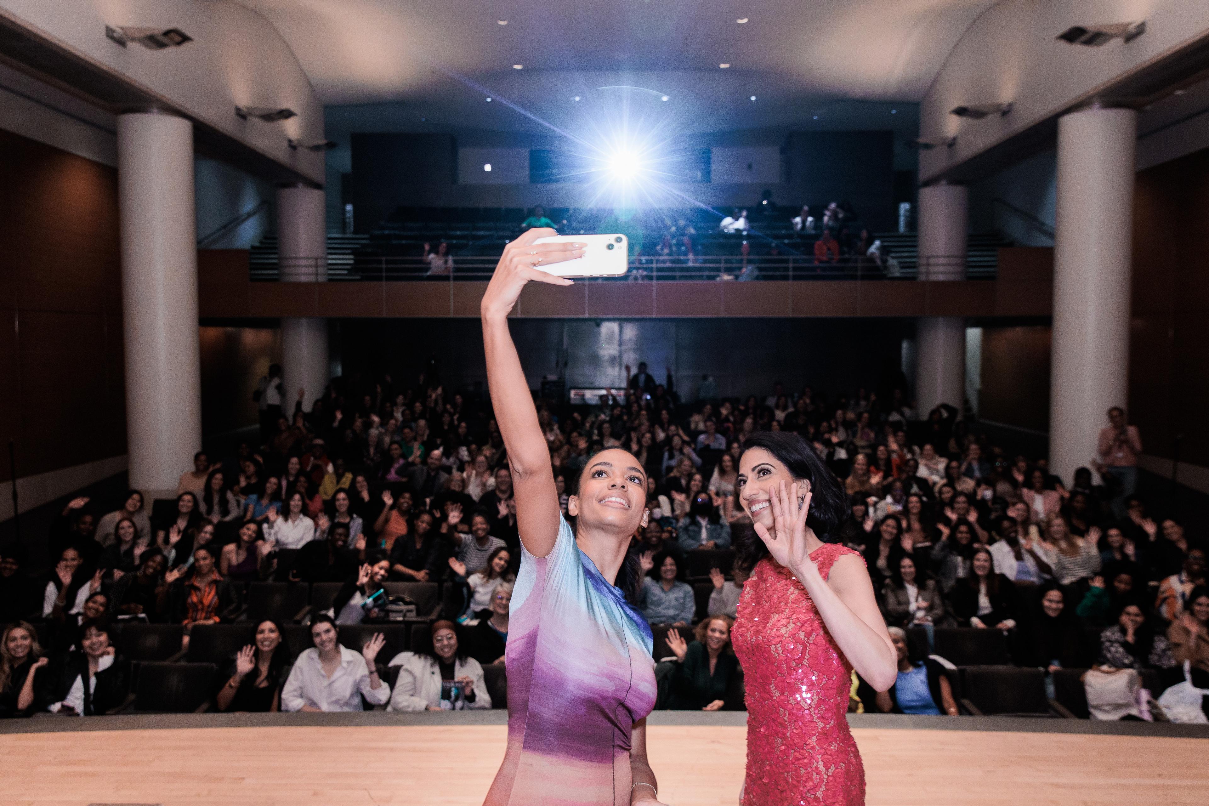 Two smiling people take a selfie on a stage with a filled auditorium behind them.