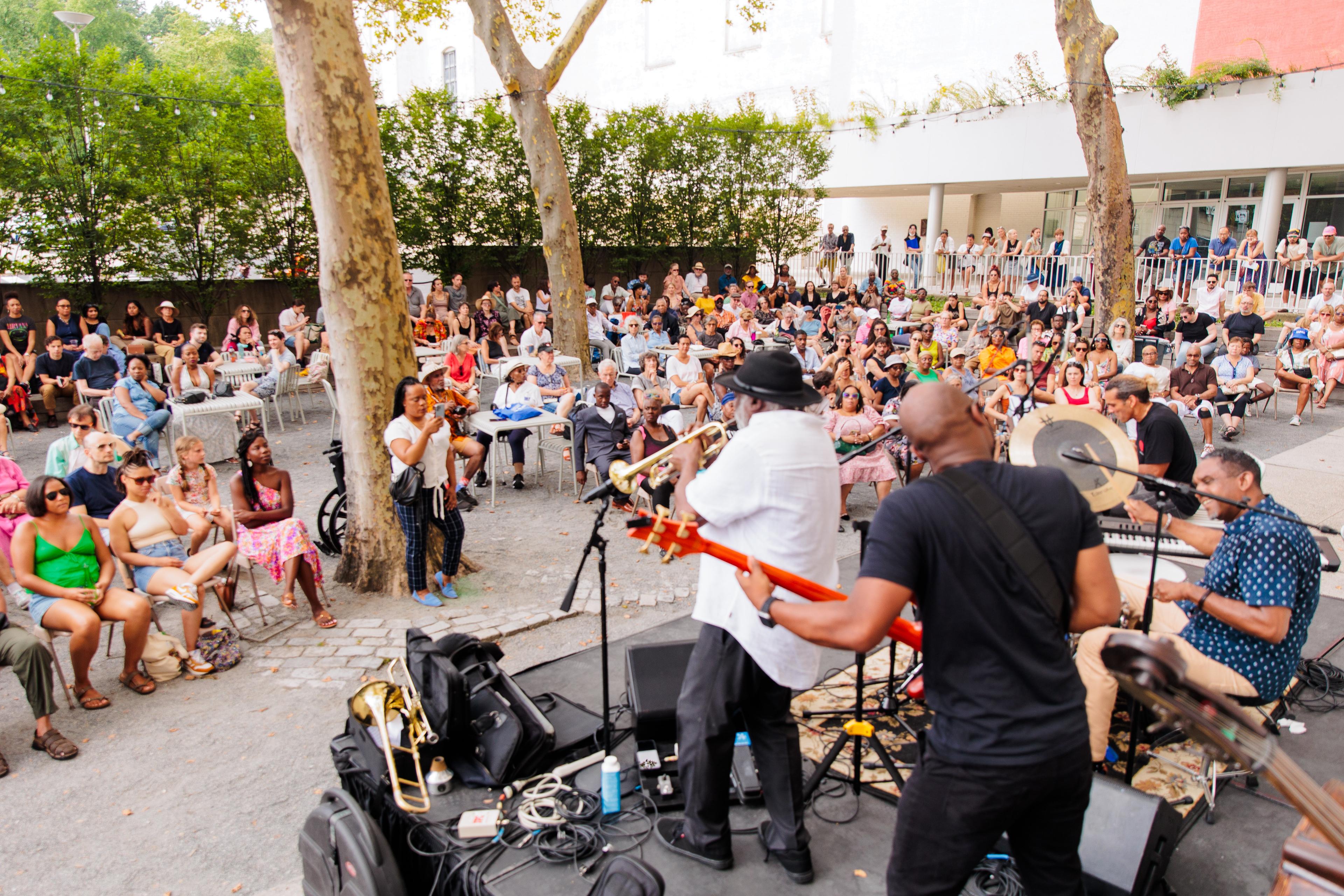A jazz band stands on an outdoor stage performing for a large audience surrounded by trees