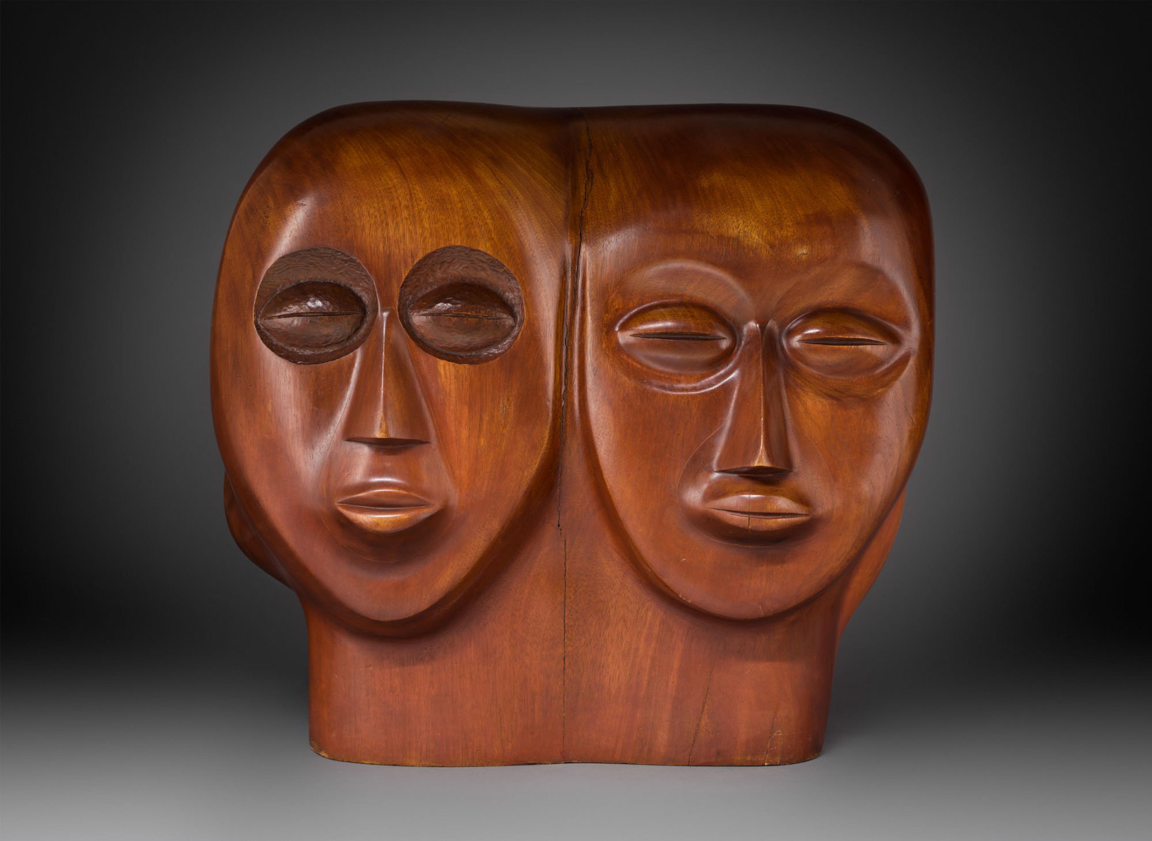 Wooden sculpture of two faces, set against a gradiant grey background