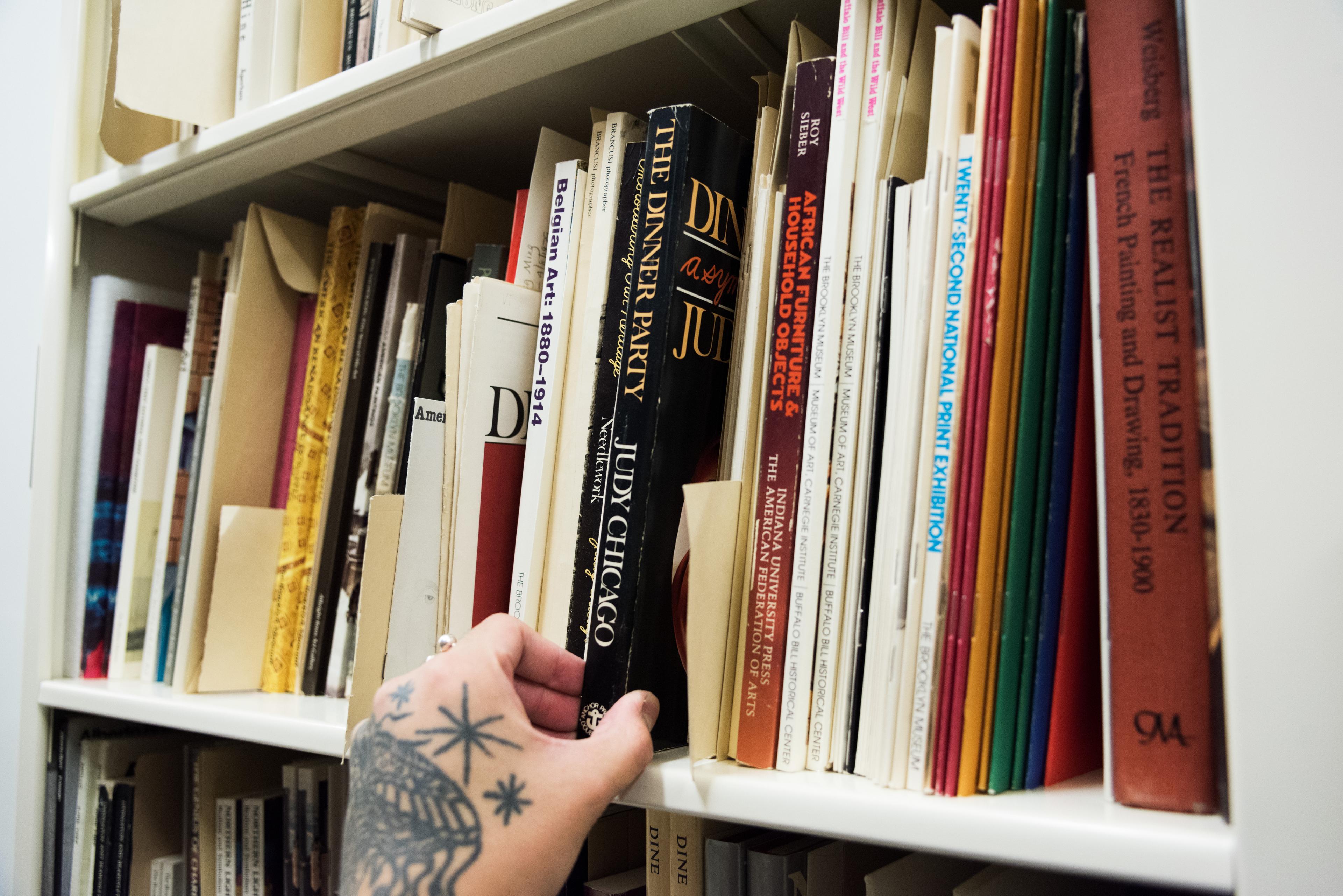 A tattooed hand pulls a book titled "The Dinner Party, Judy Chicago" from a bookshelf.