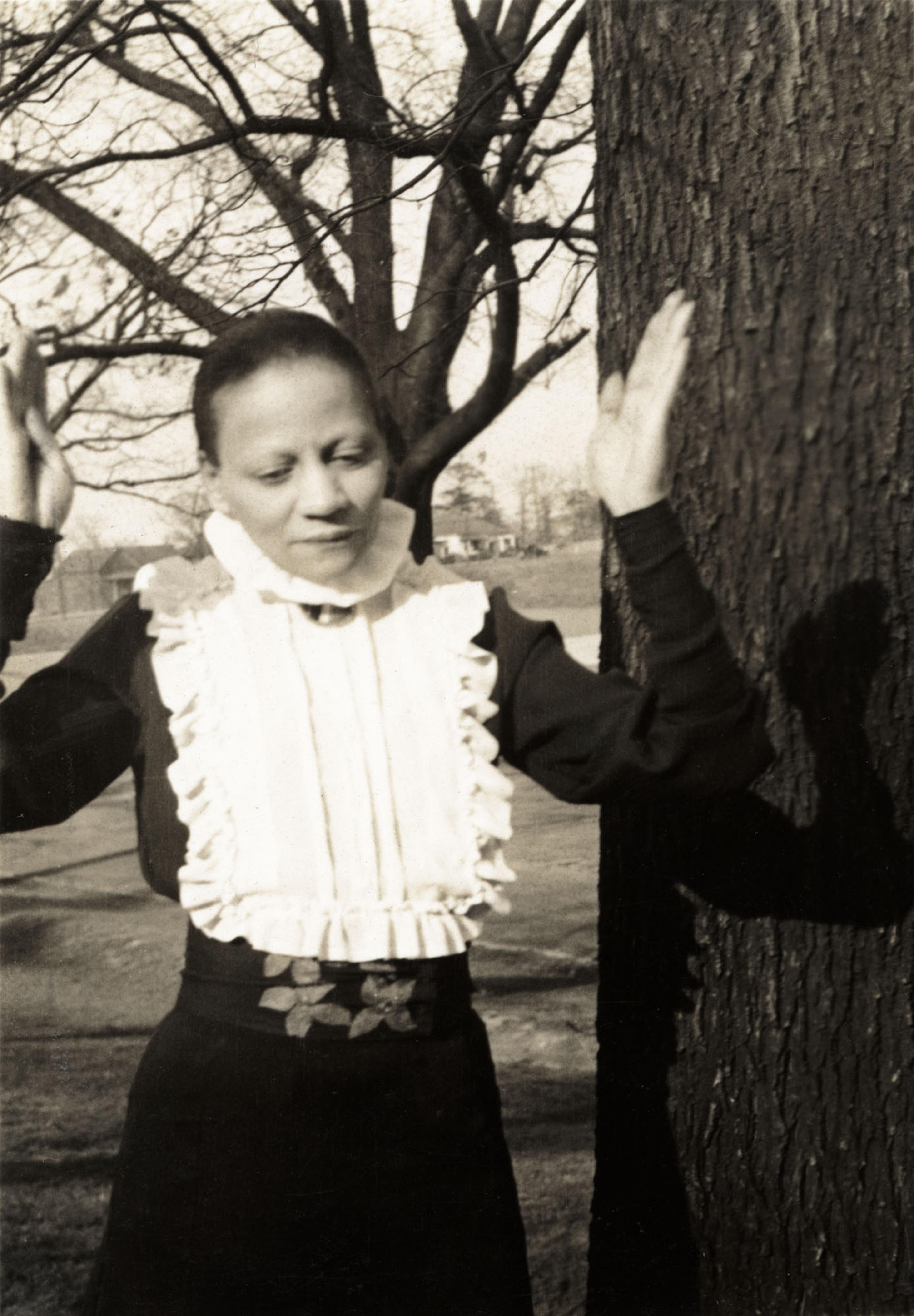 Black-and-white photograph of a woman raising her hands while standing beside a tree