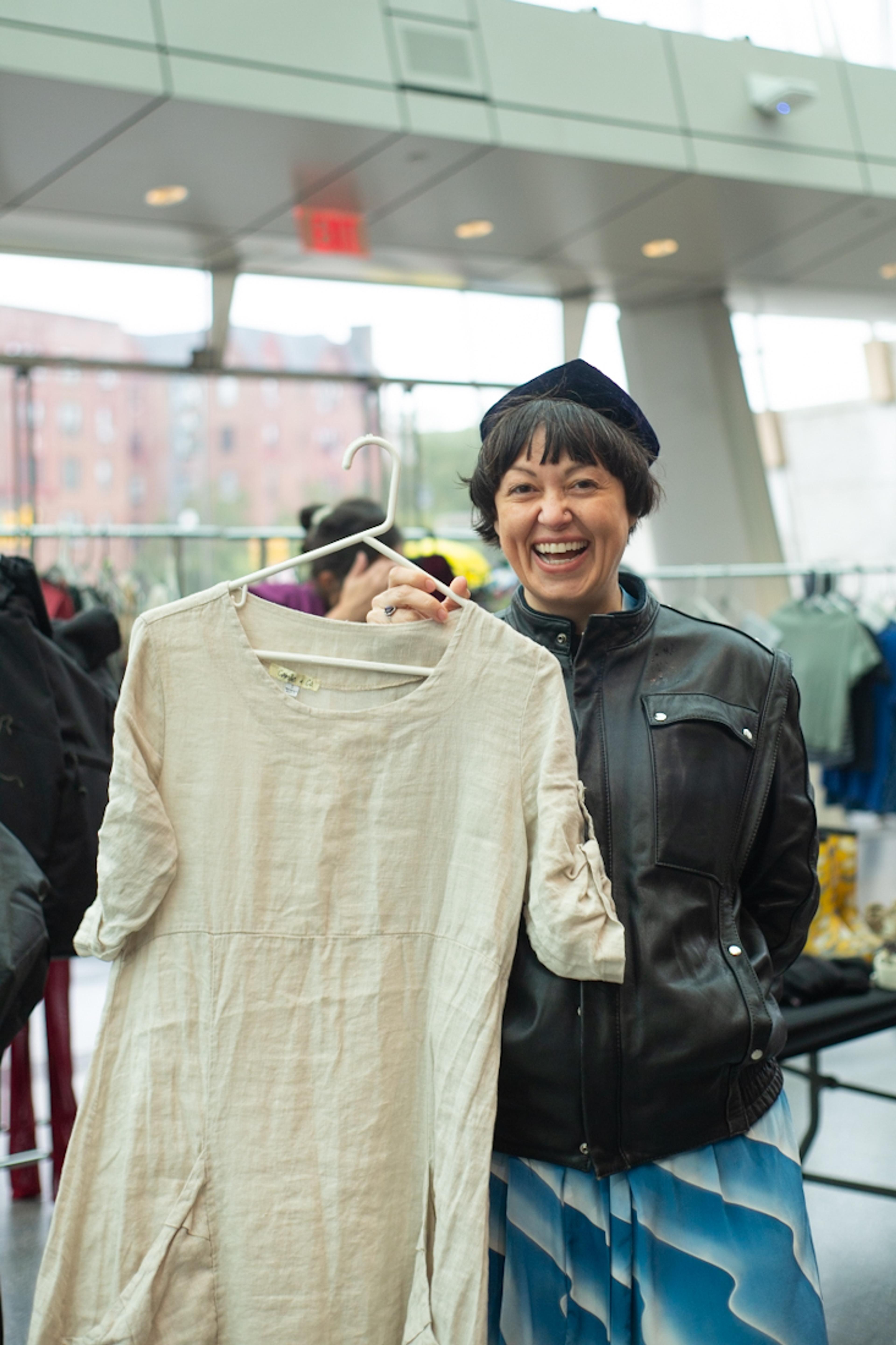 Person in a black hat and black leather jacket holds a cream colored dress on a hanger smailing