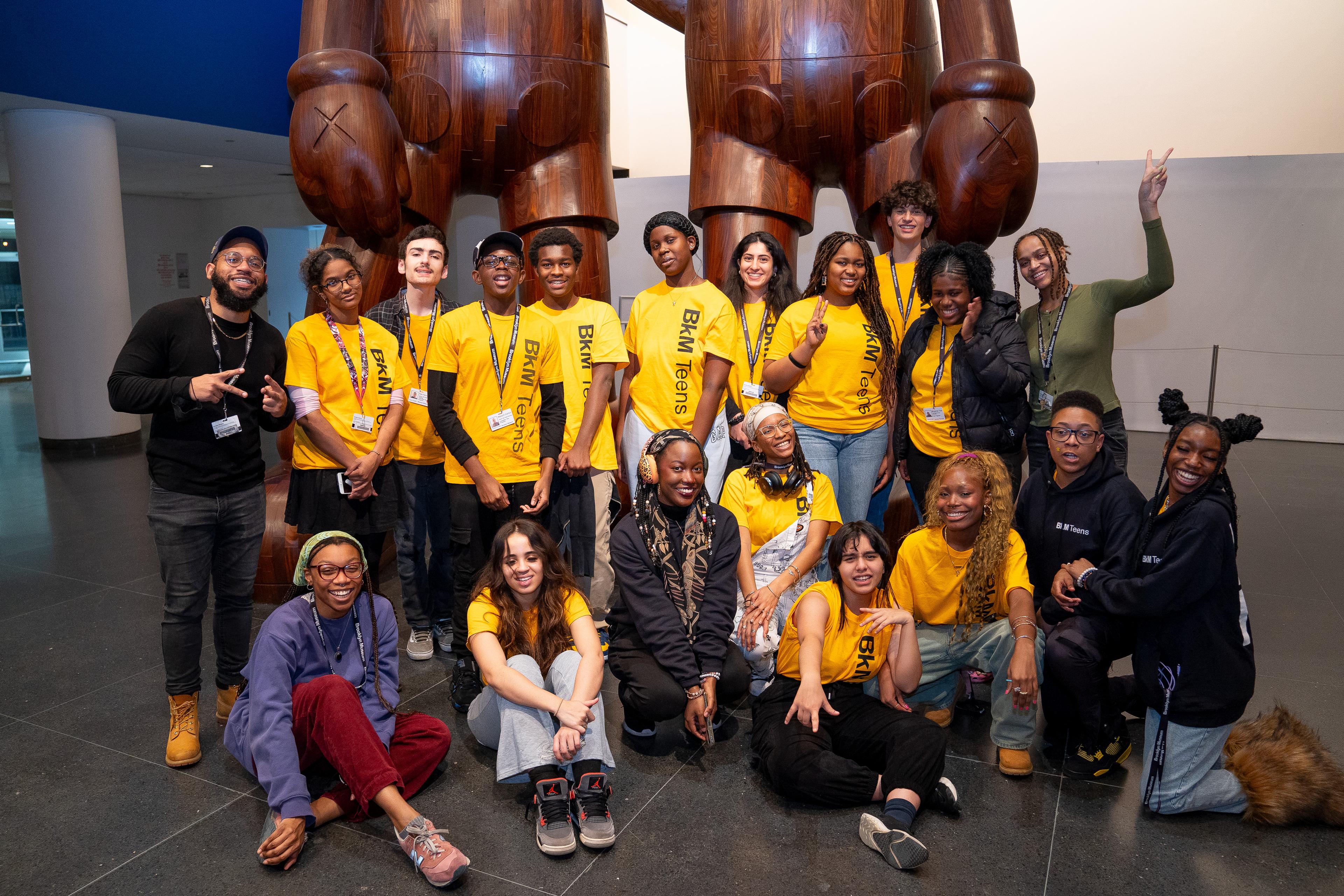 Group of teens wearing yellow "BKM Teens" shirts pose smiling in front of a tall wooden sculpture by KAWS.