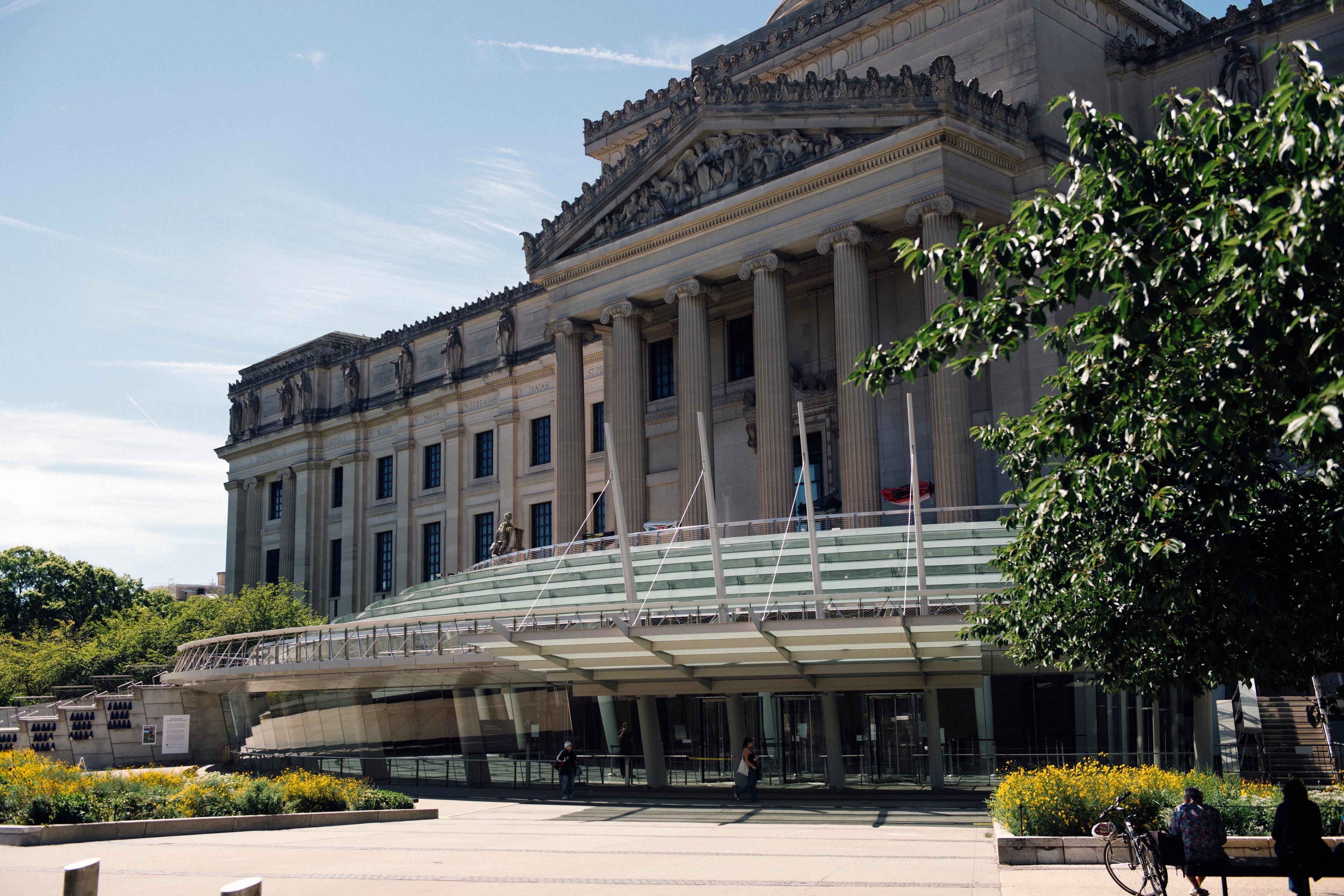 People sitting on shady benches on a green plaza in front of a large neoclassical building with a modern glass entrance