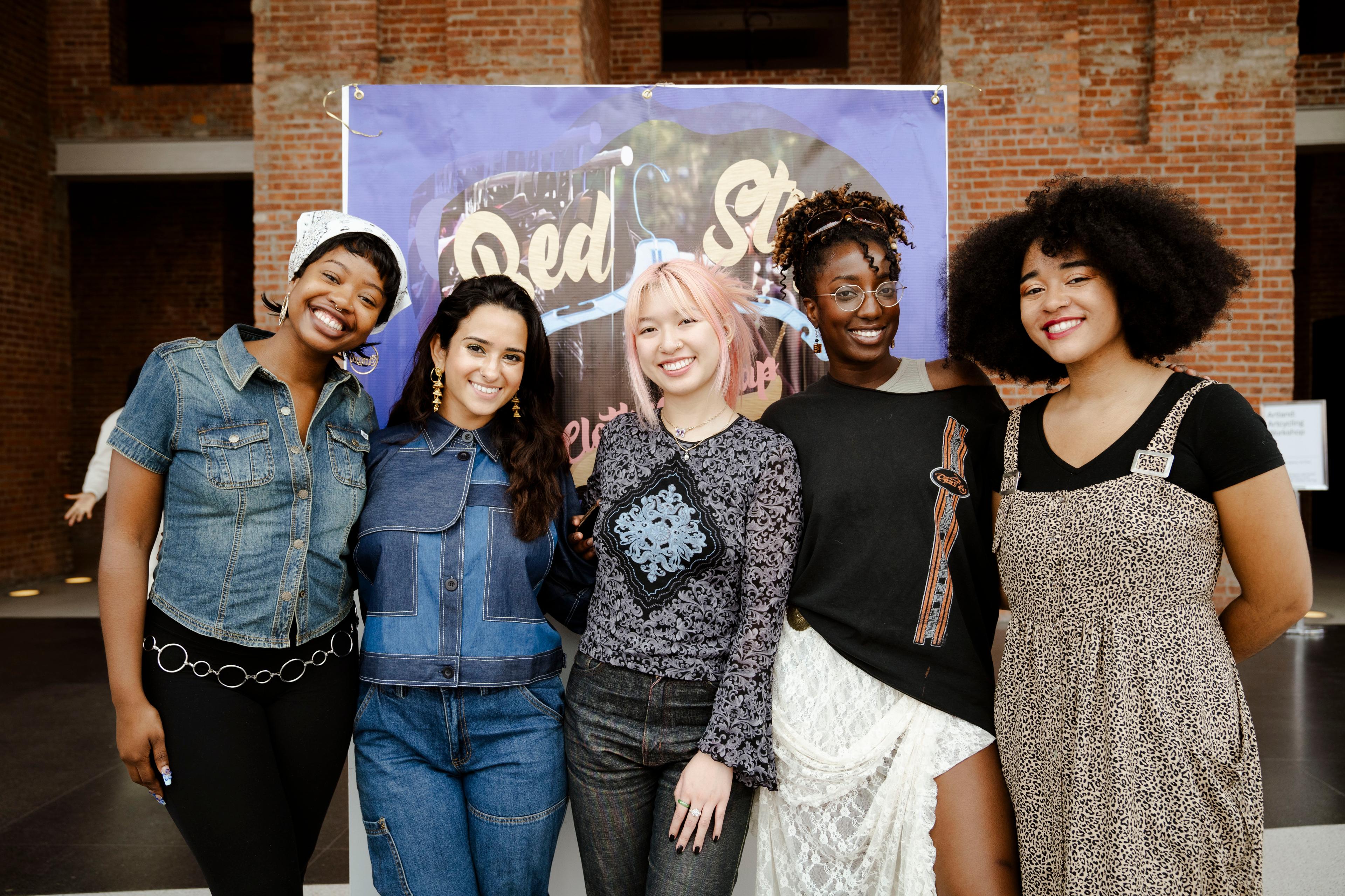 Group of five adults pose together smiling in front of a backdrop that reads "Bed-Stuy Clothing Swap"