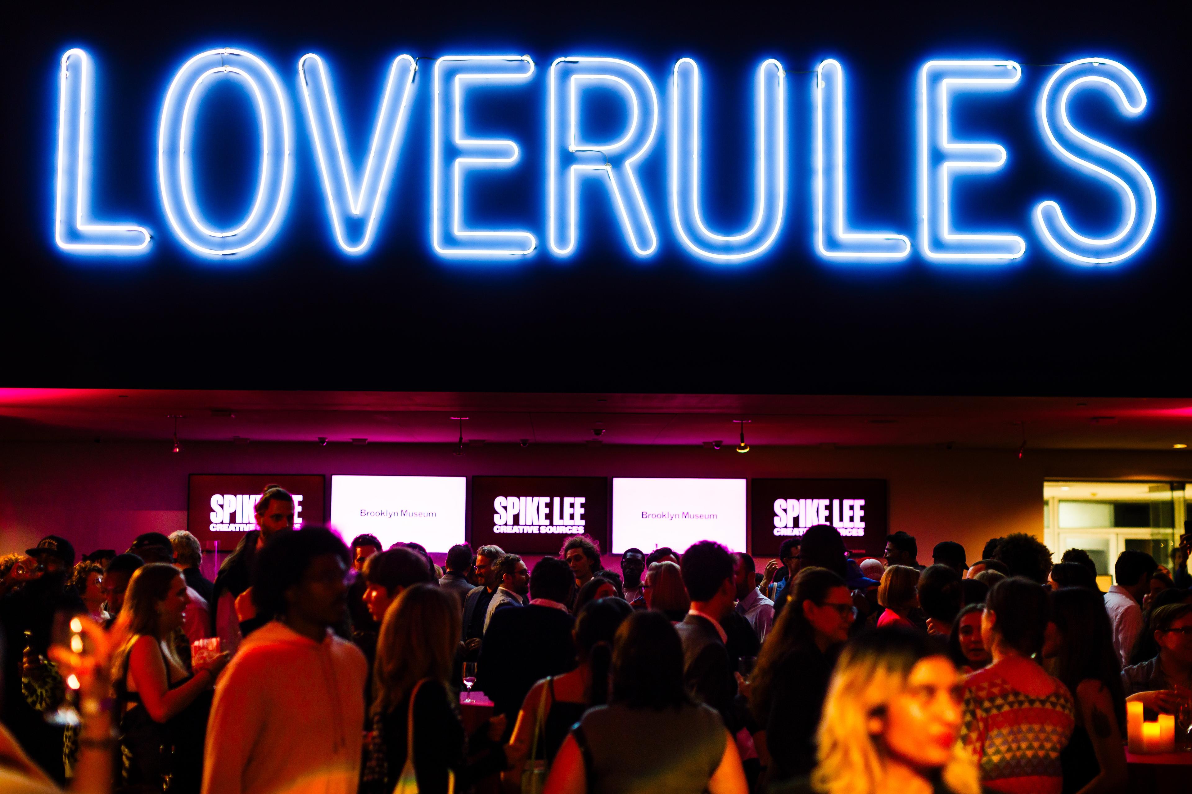 Party goers fill a large, dark room with a large neon artwork above that reads "LOVERULES"