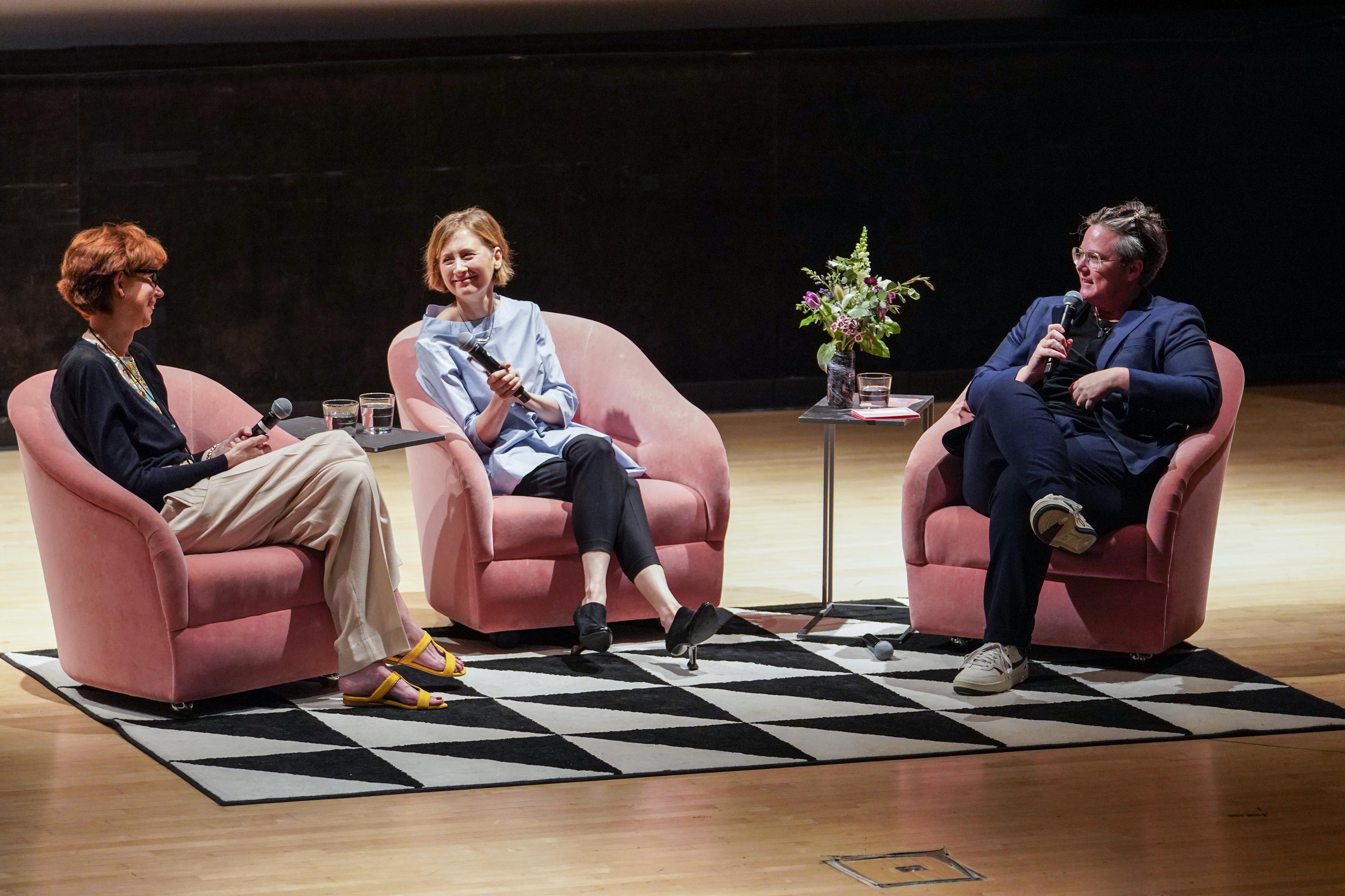 Three people holding mics sit on a stage in pink chairs on top of a black and white graphic rug