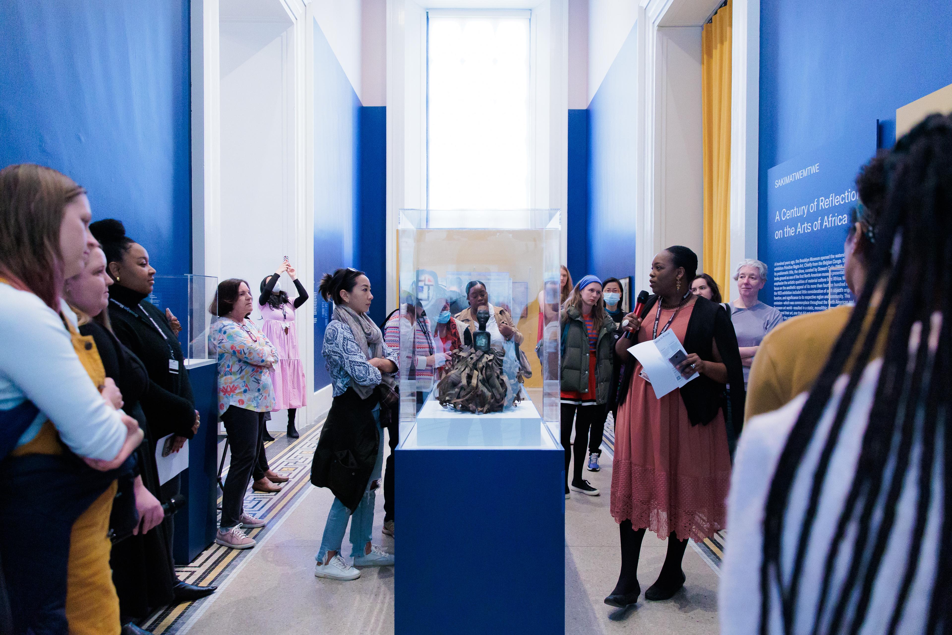 A group of visitors surround a person speaking into a microphone next to a sculpture