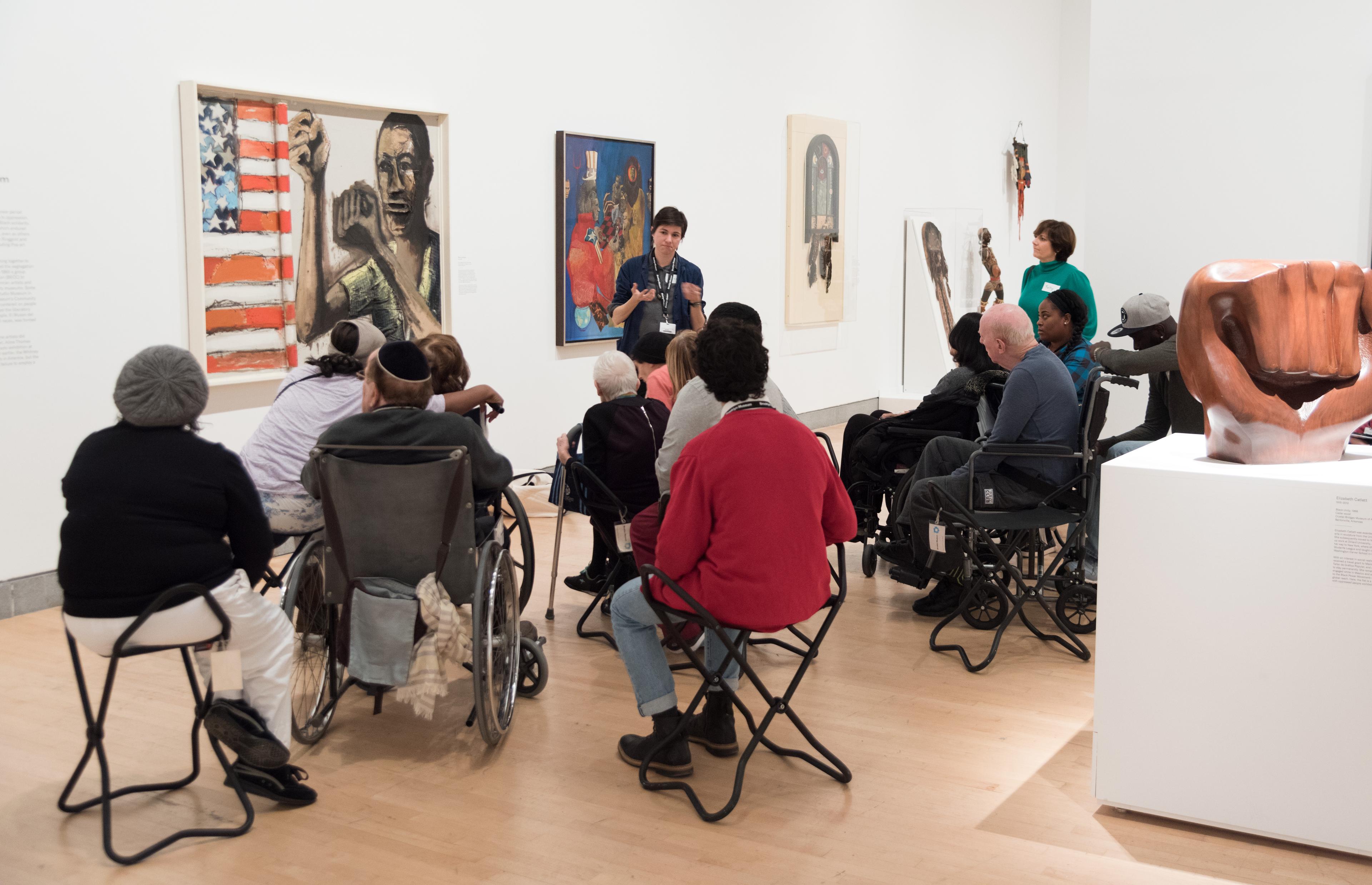 A group of adults sitting around in folding stools and wheelchairs face a painting on a wall while an instuctor with speaks to them