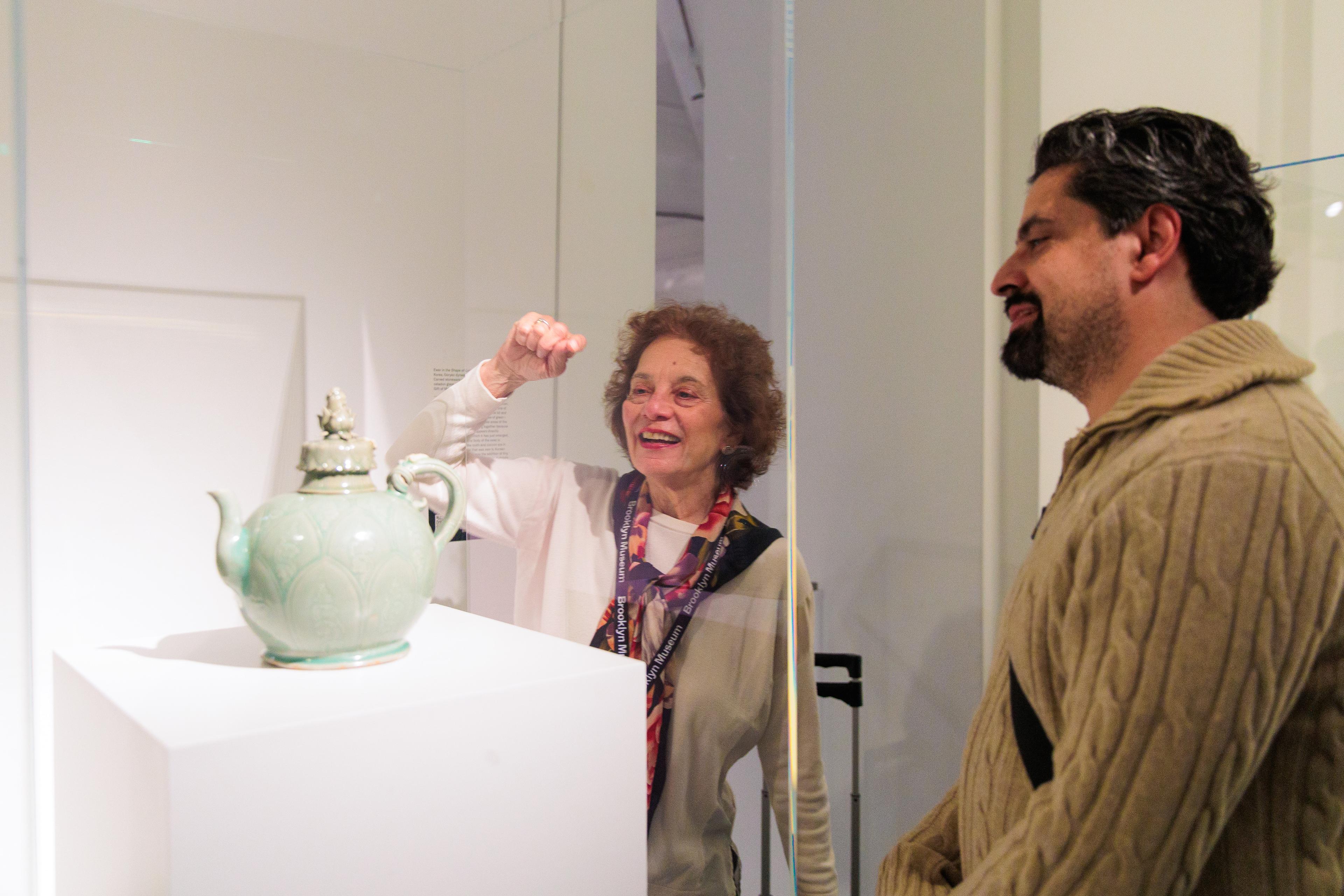 An adult speaks and gestures towards a ceramic teapot with another adult smiling on