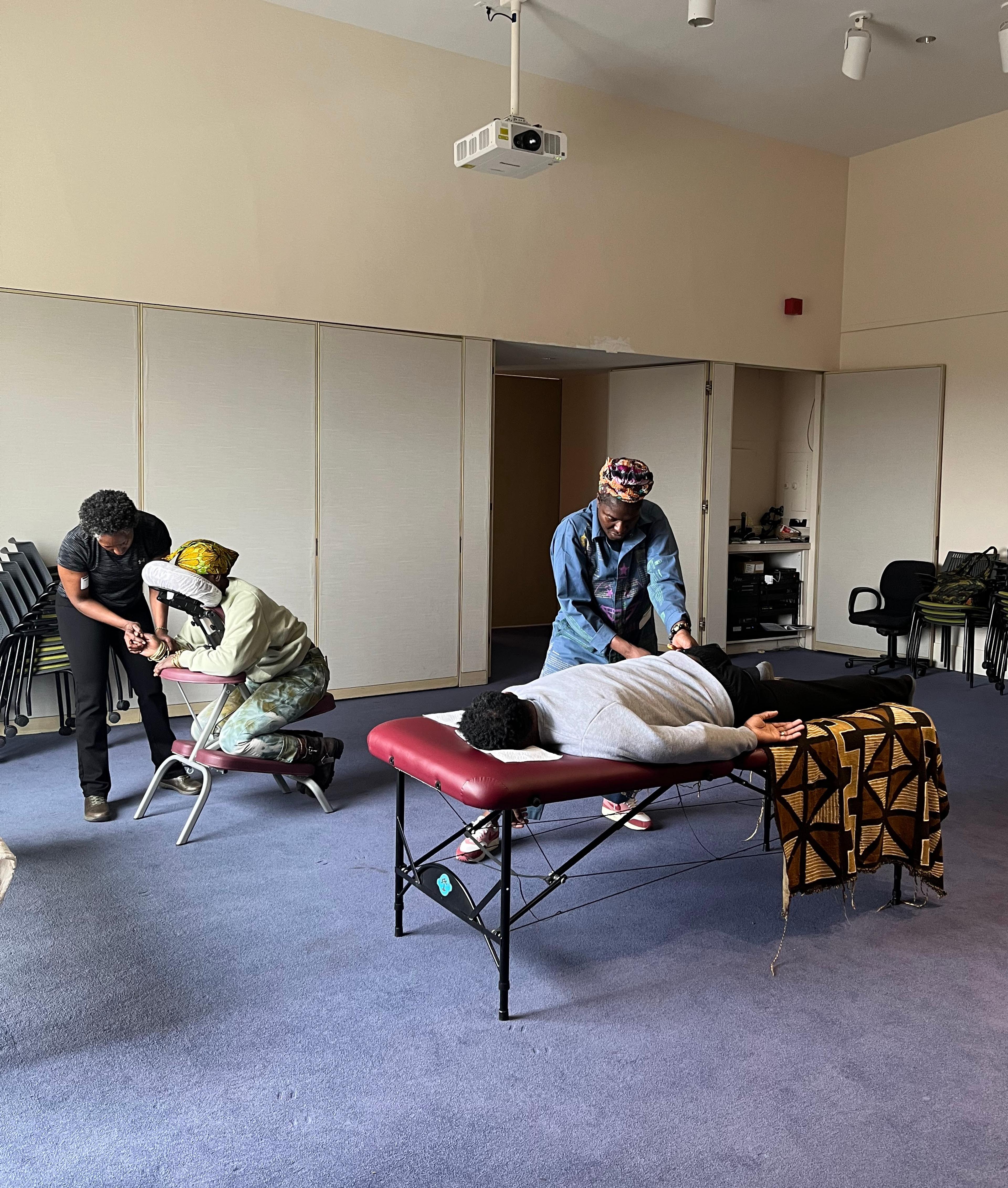 Two adults sitting and laying in a masseuse chair and table, receive massages from two other adults.