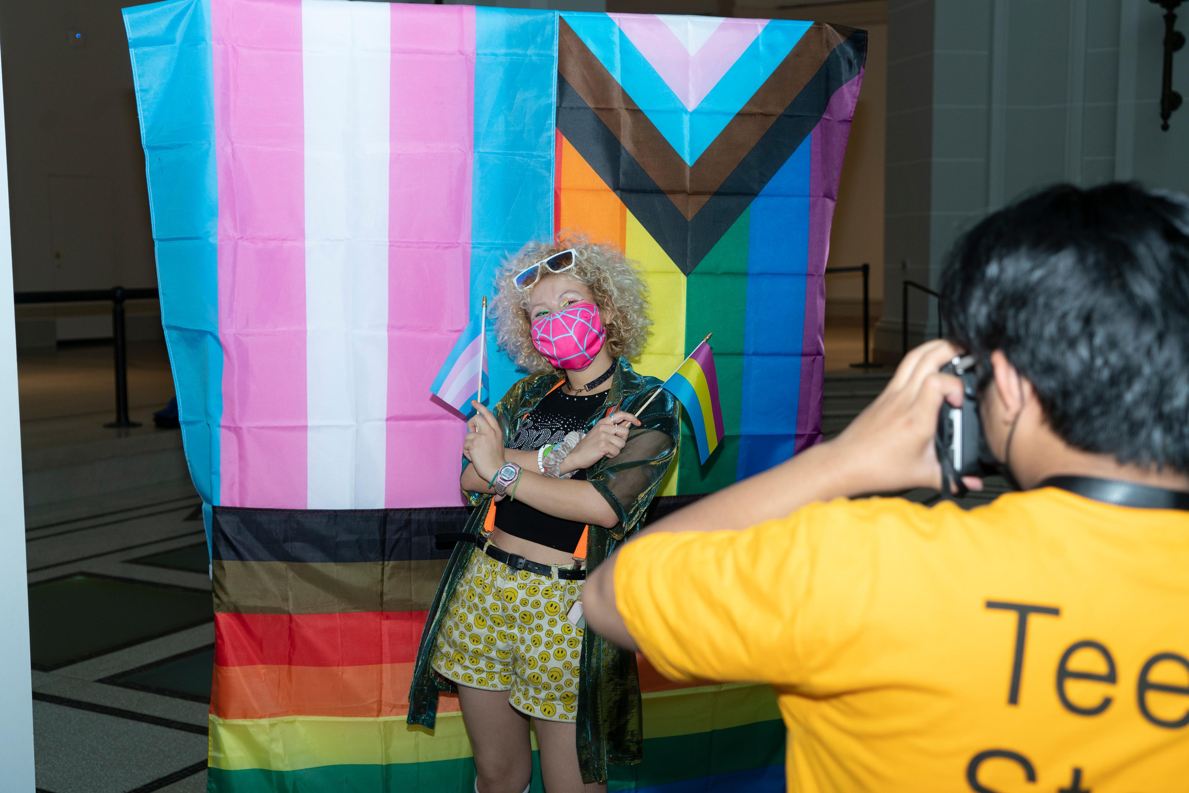 A blonde teen poses in front of LGBTQ+ pride flags holding two small Trans and Pansexual pride flags while another teen takes their photo