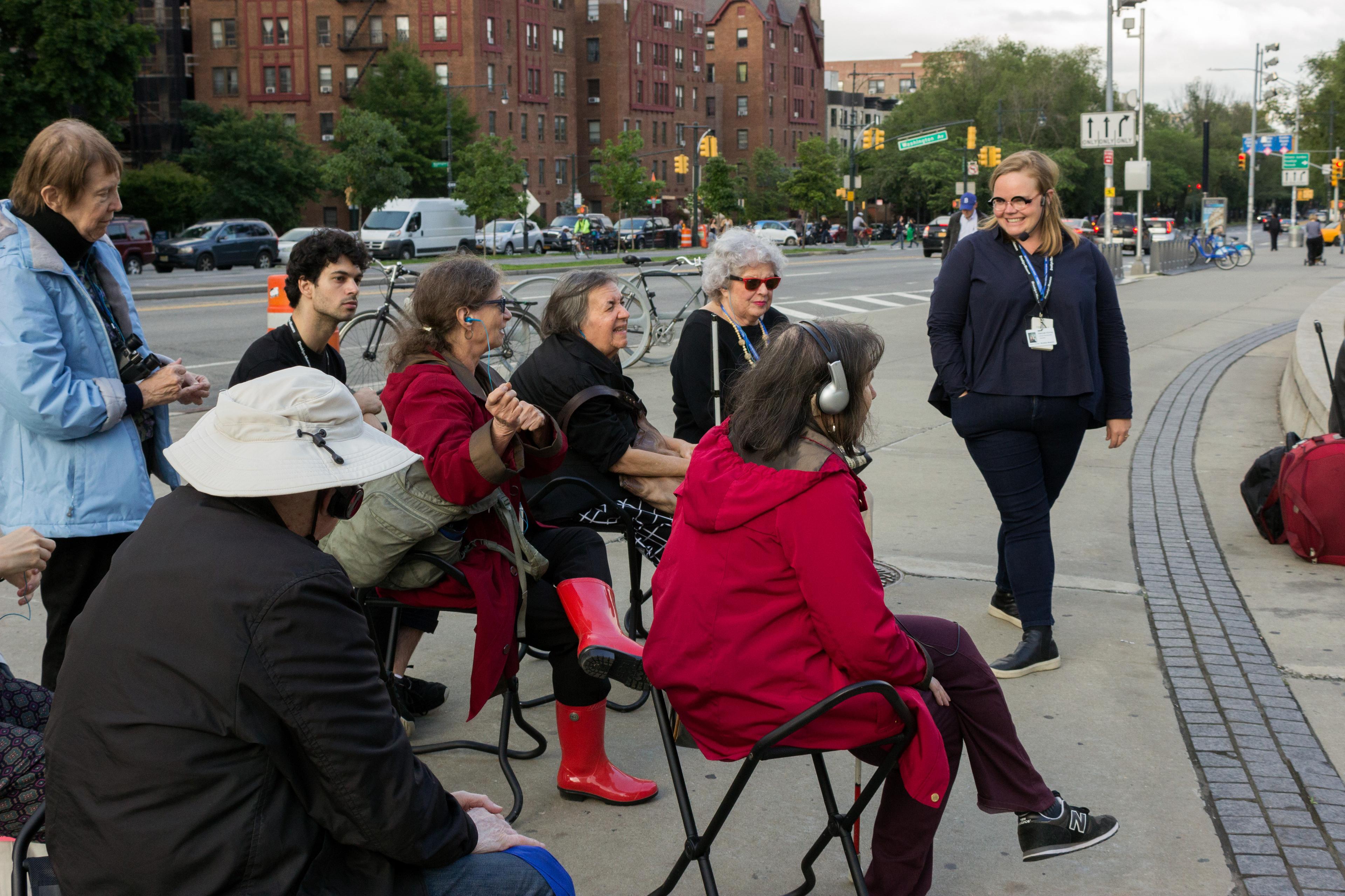 A group of adults wearing headphones and sunglasses sit outside on folded stools speaking with a standing instructor wearing a mic