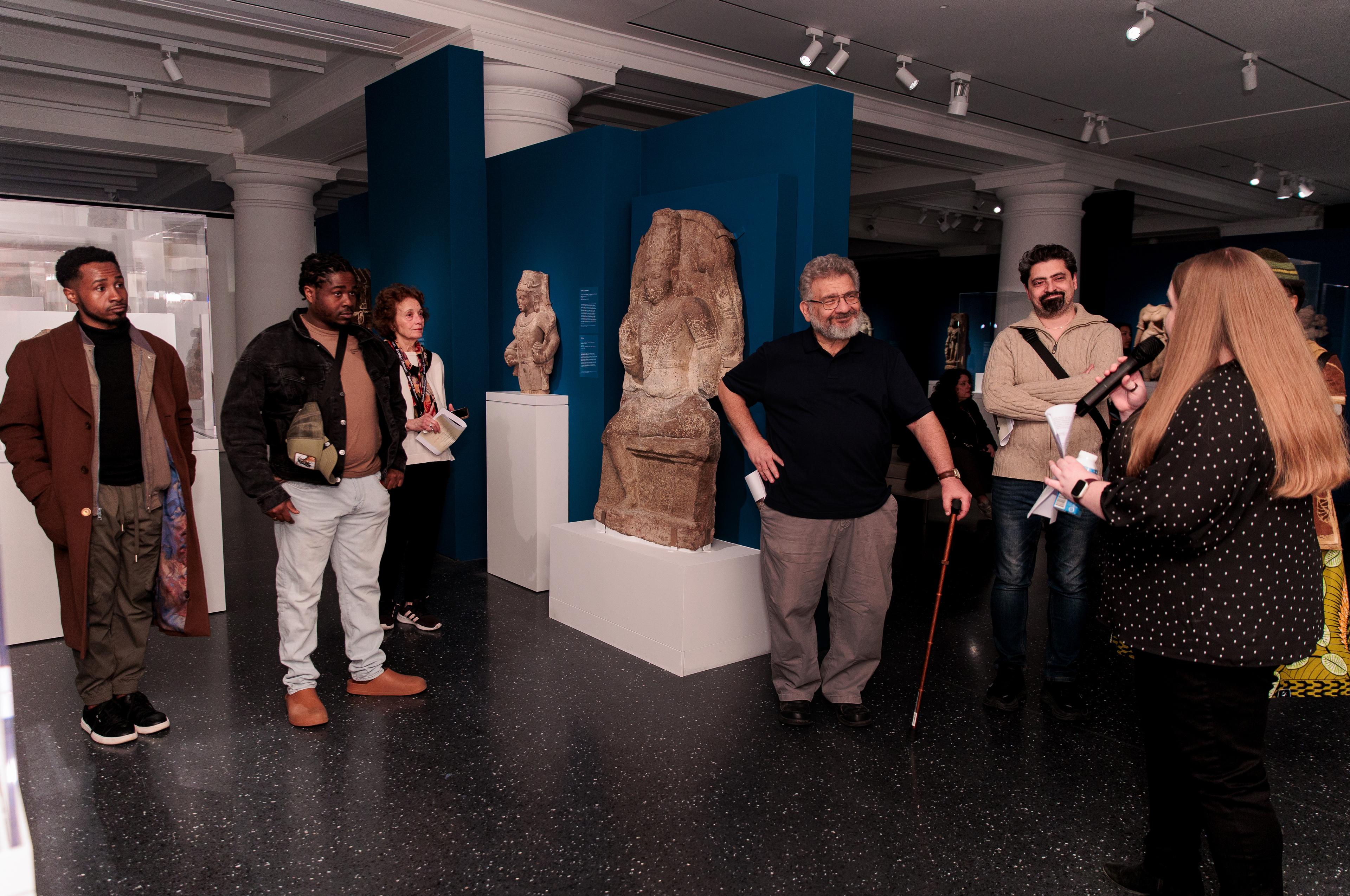 A person with a microphone addresses a group of adults standing in an art gallery