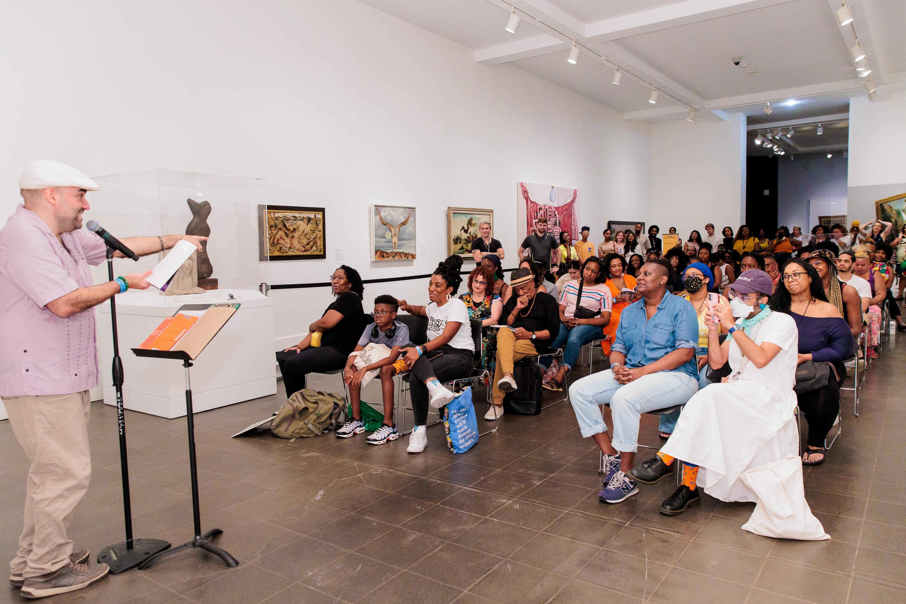 Dozens of people sit inside an art gallery facing a spoken word performer smiling back