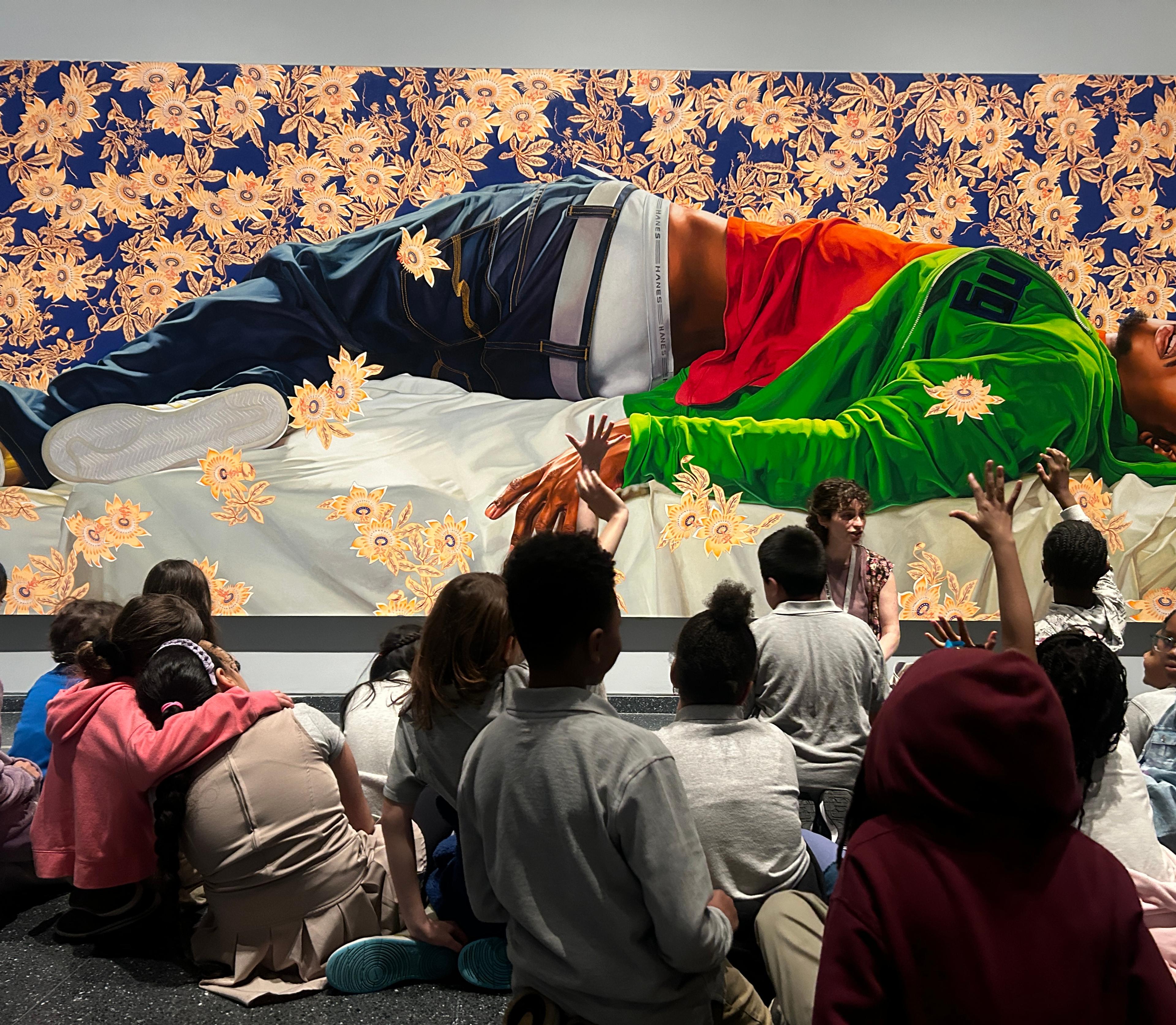 A young adult squats infront of a large painting of a reclining figure, speaking to a group of young students