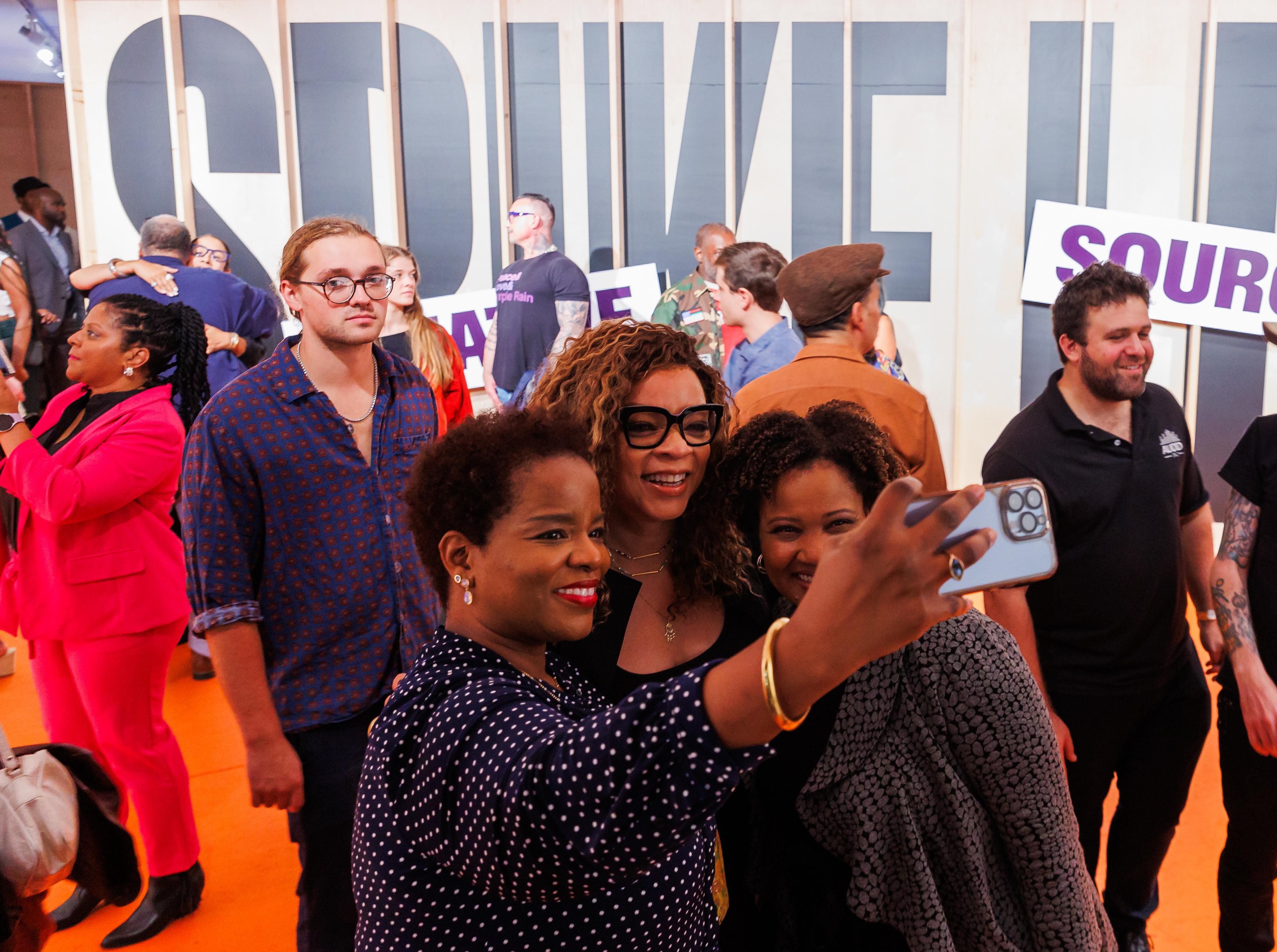 A group of three people pose for a selfie in front of an exhibition gallery wall that reads "Spike Lee Creative Sources"