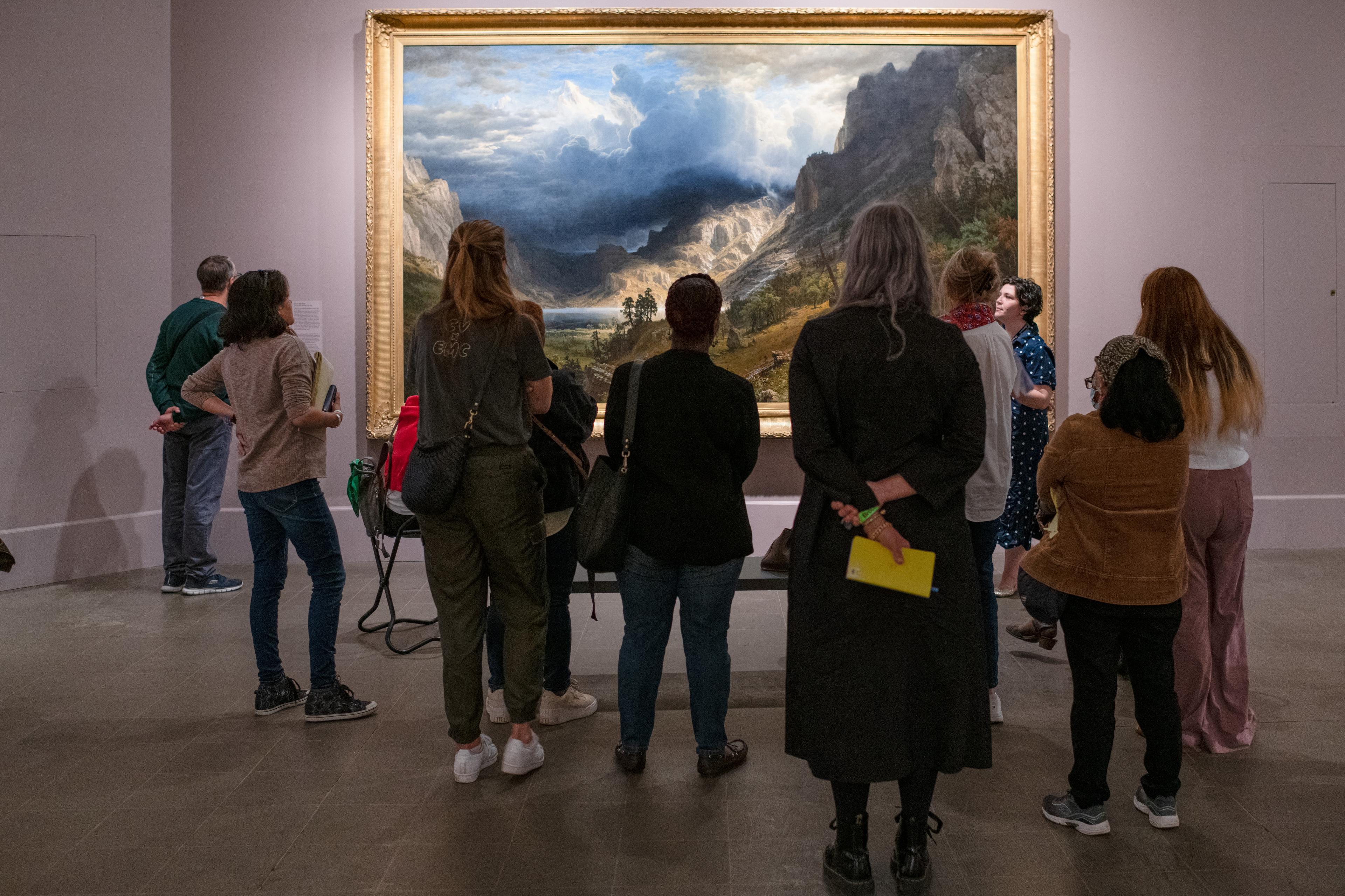 A group of adults all face a large landscape painting while an educator speaks