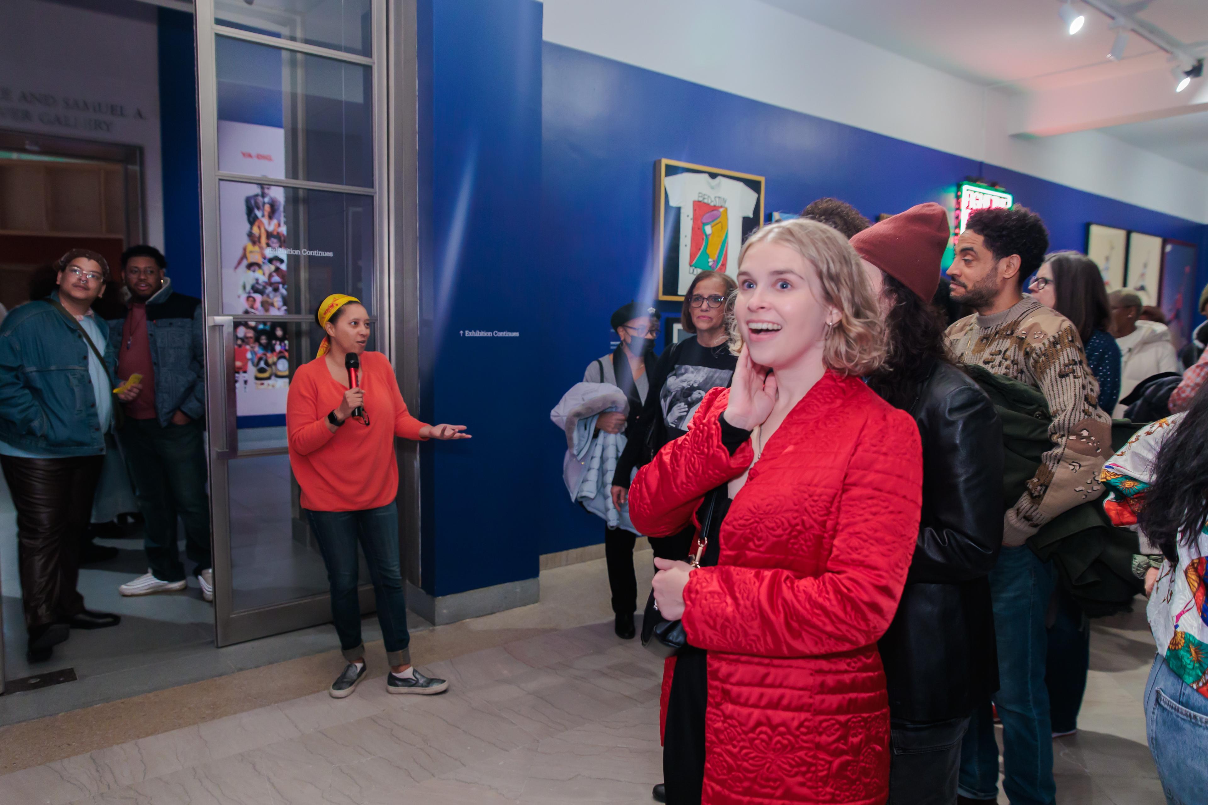 A curator in a orange sweater and yellow head wrap speaks and gestures to a group of happy and excited people in a blue gallery full of objects