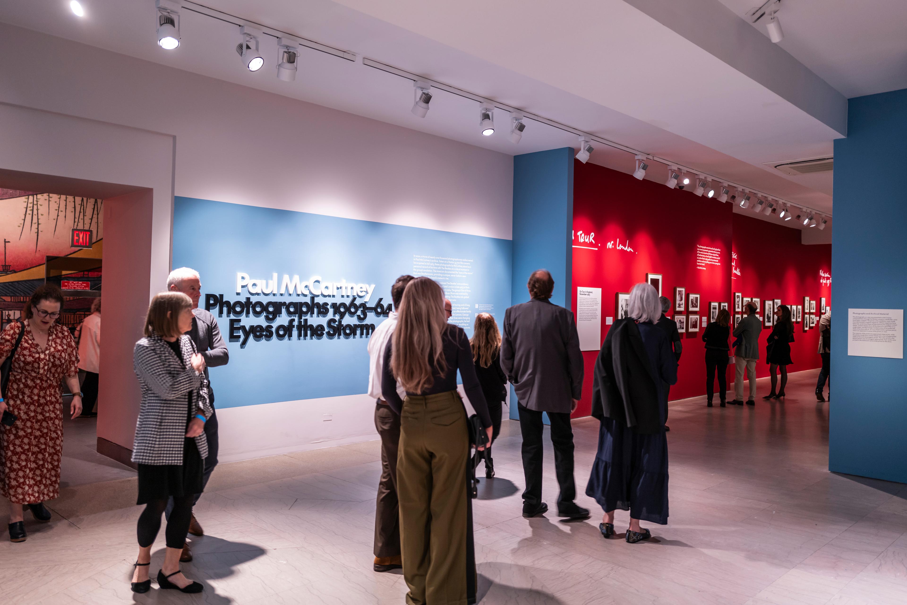 A group of visitors walk into an exhibition gallery, passing a wall that reads "Paul McCartney Photographs 1963–64 Eyes of the Storm"