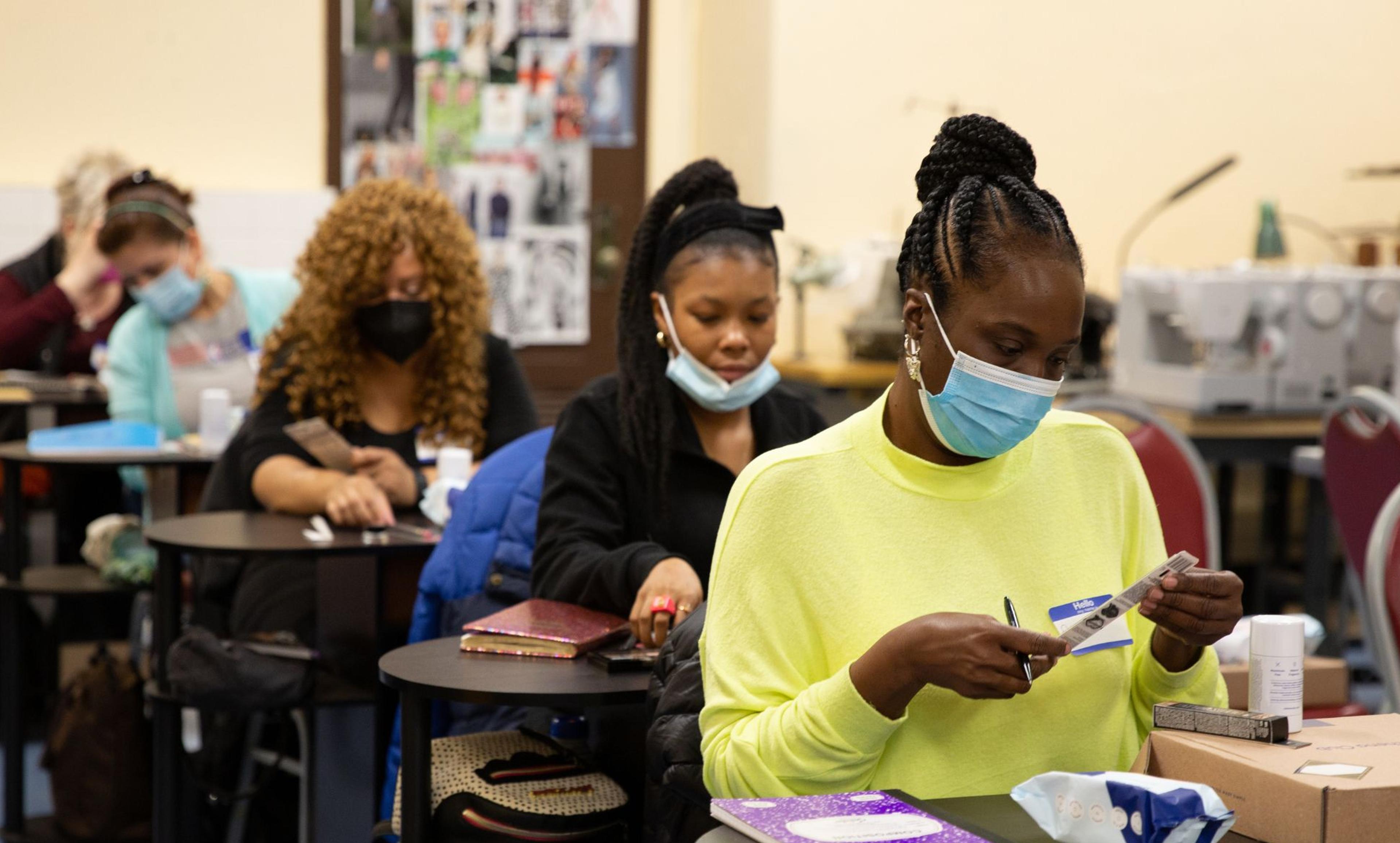 Four adults sit in a row of desks handling sewing materials