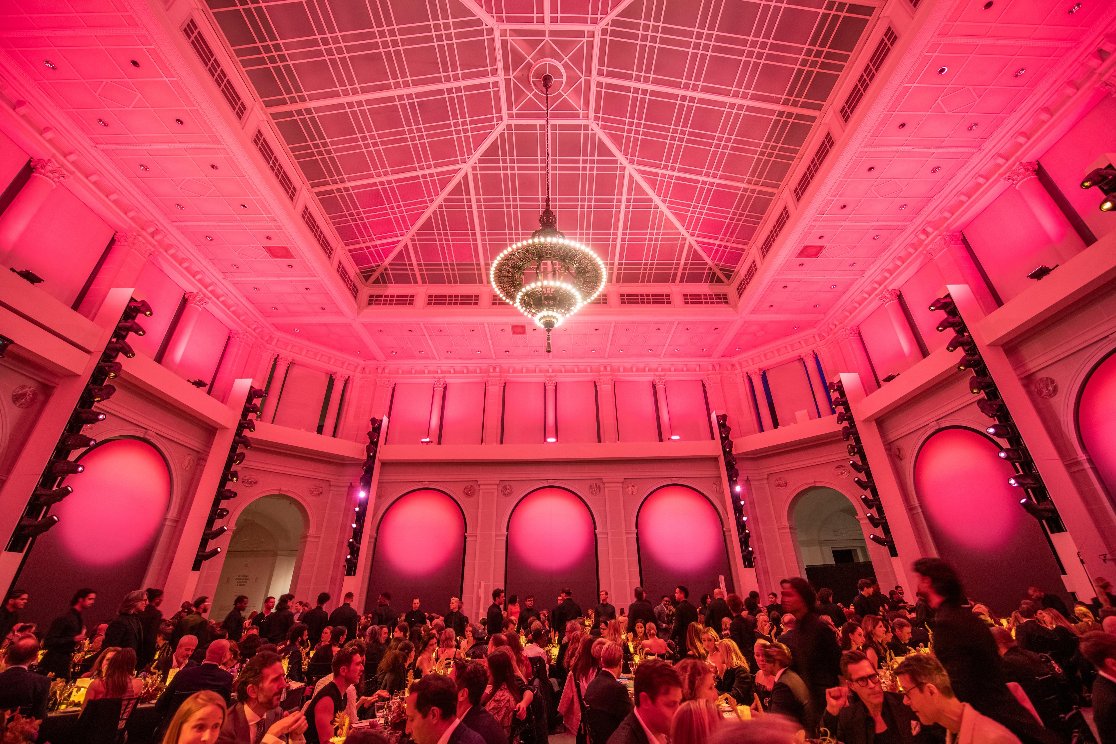 A room full of guests seated at tables fill a large ball room with a chandelier and pink lighting