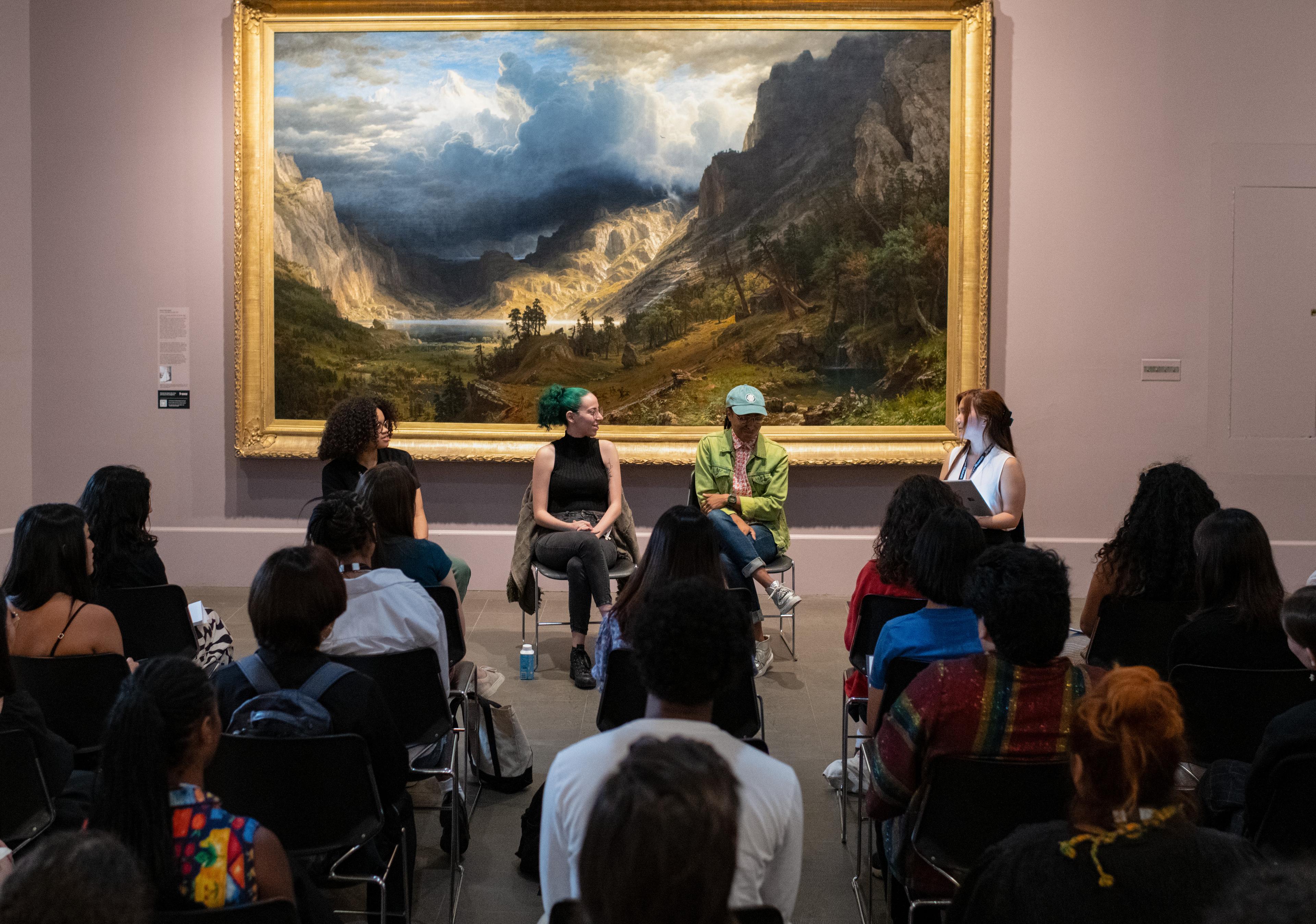 A group of seated adults face four panelists speaking in front of a large painting