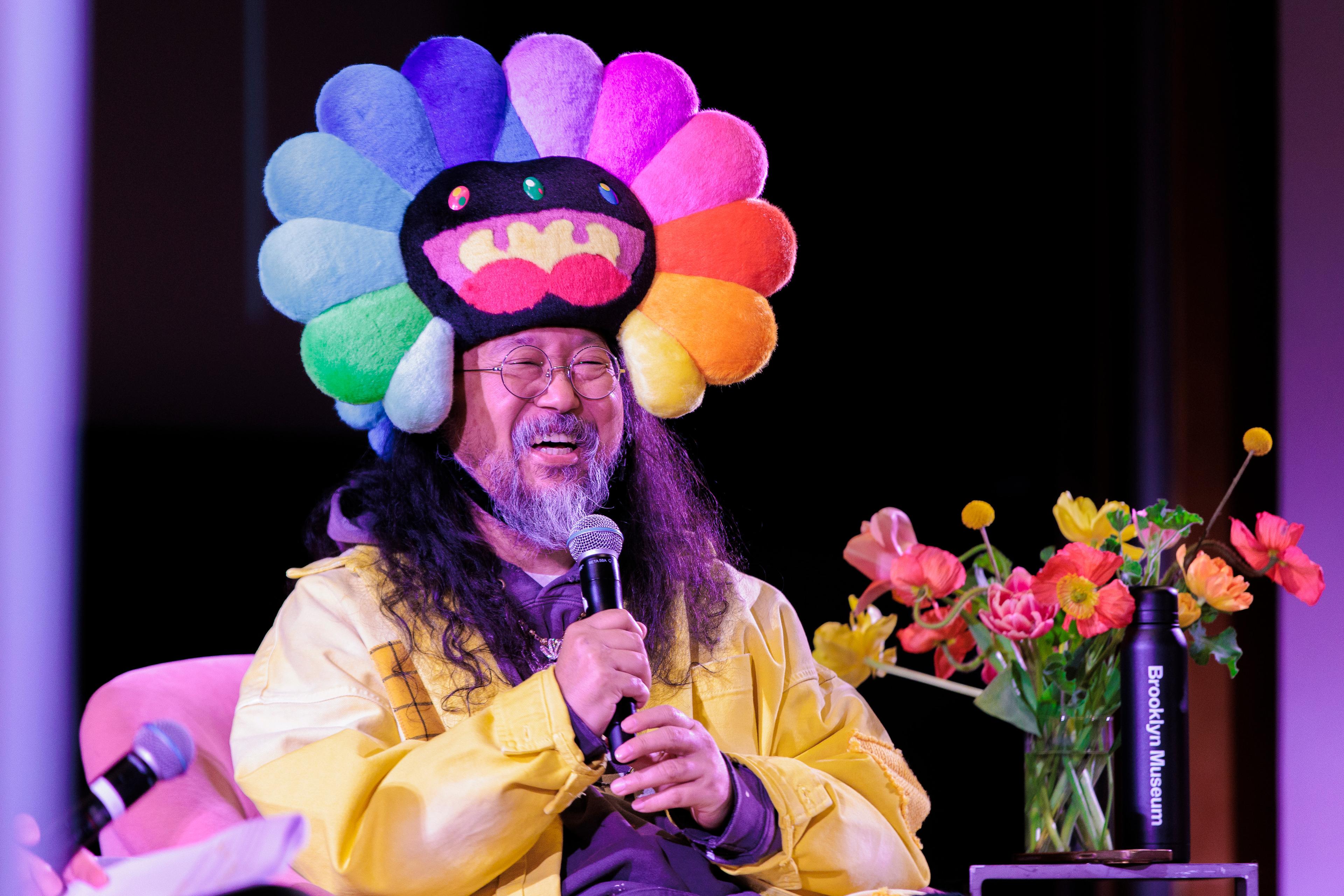 Smiling person in a multi-colored flowered hat, holds a mic while laughing