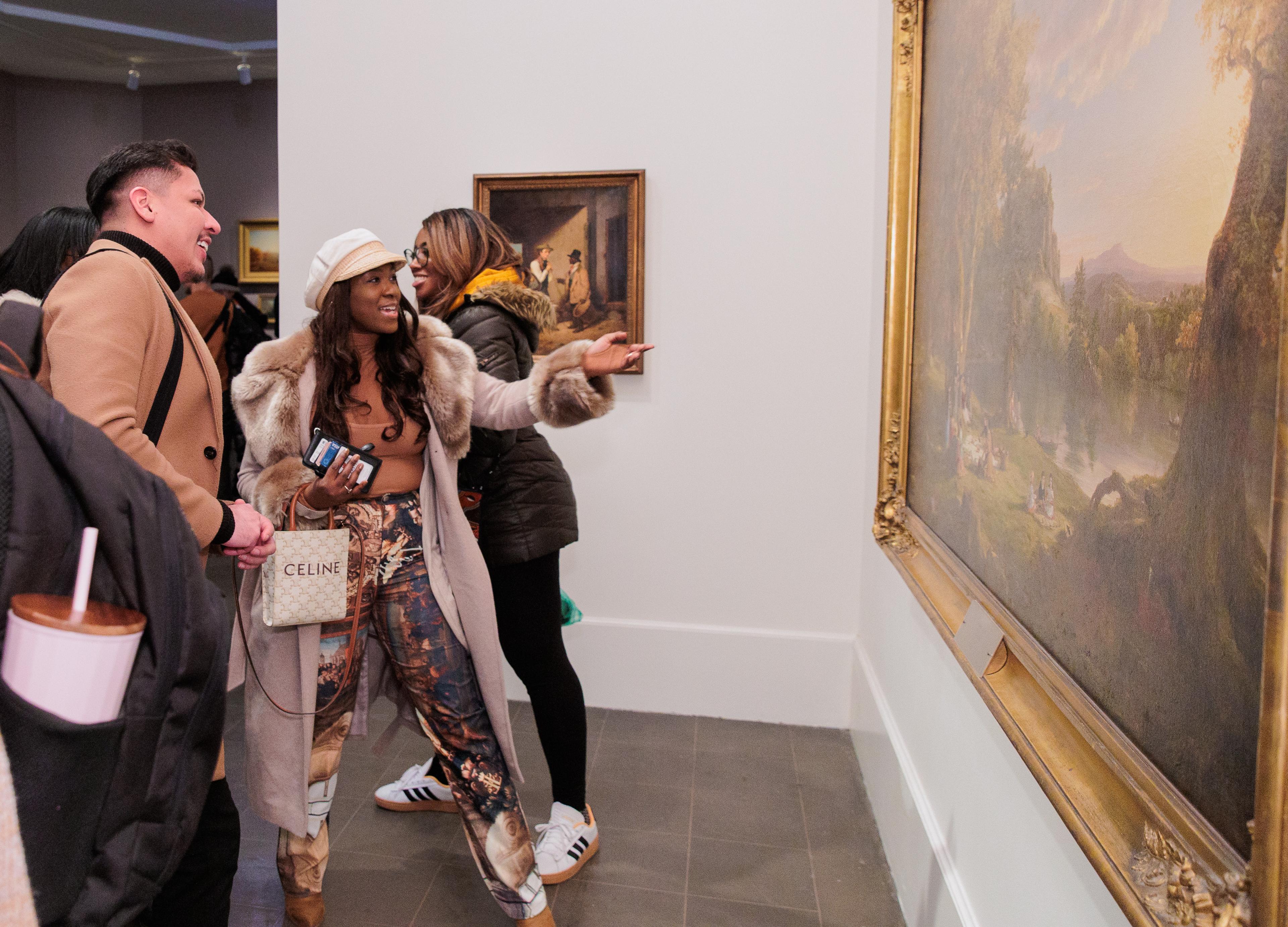 A couple friends in a busy gallery smile and gesture towards a large landscape painting