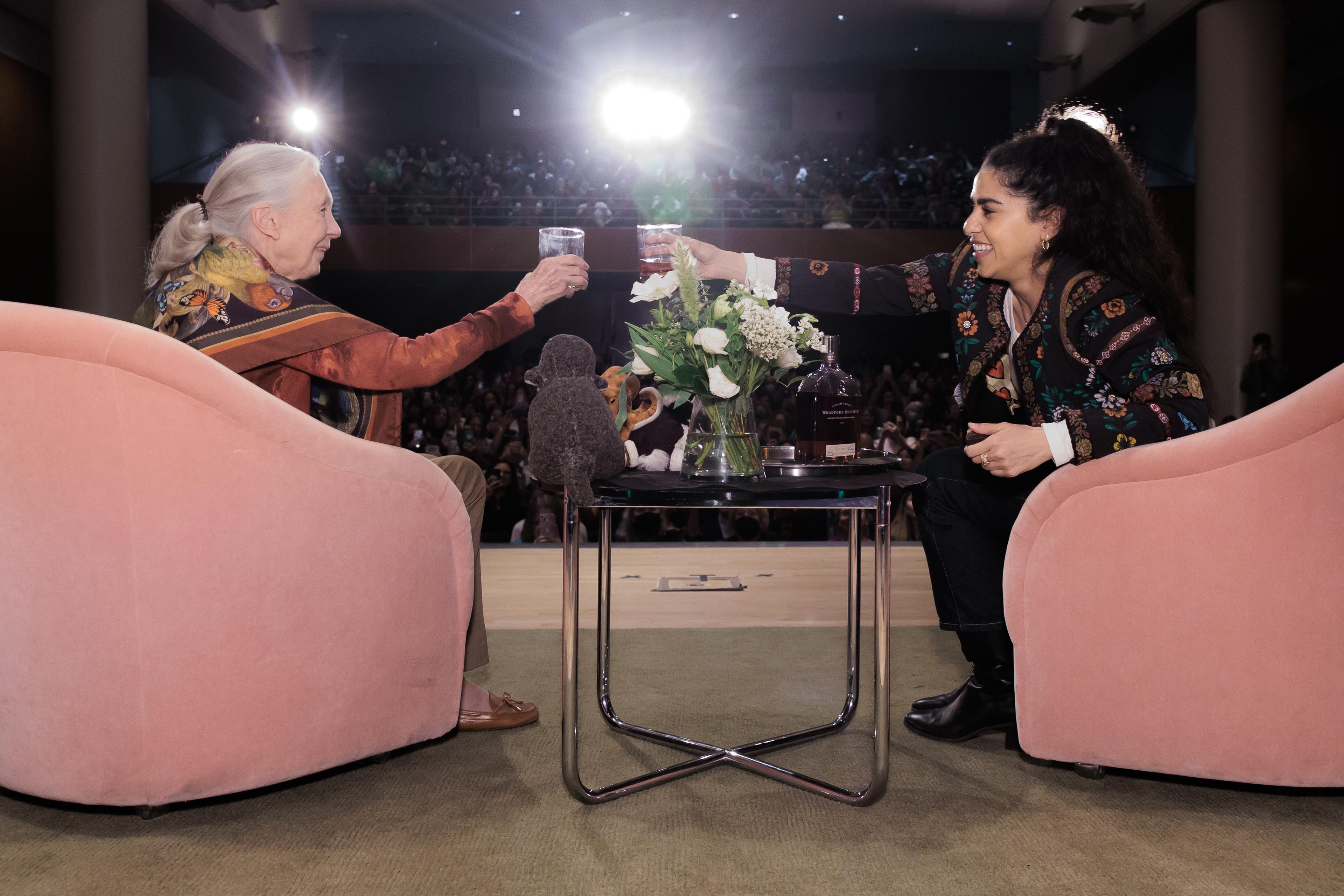 Two people seated in pink chairs toast and smile at eachother onstage with a crowded auditorium in the background