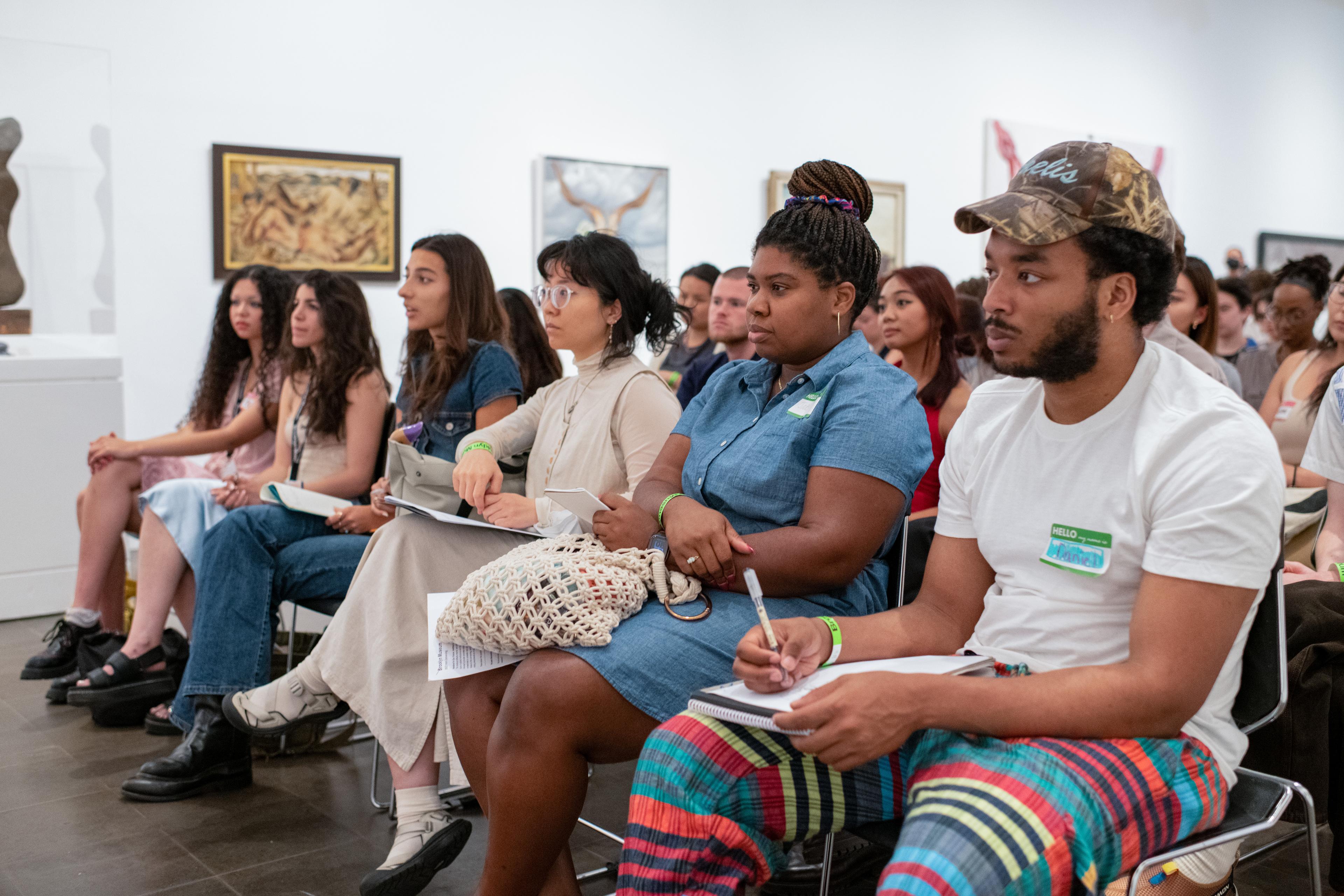 A large group of seated adults listen attentively in an art gallery