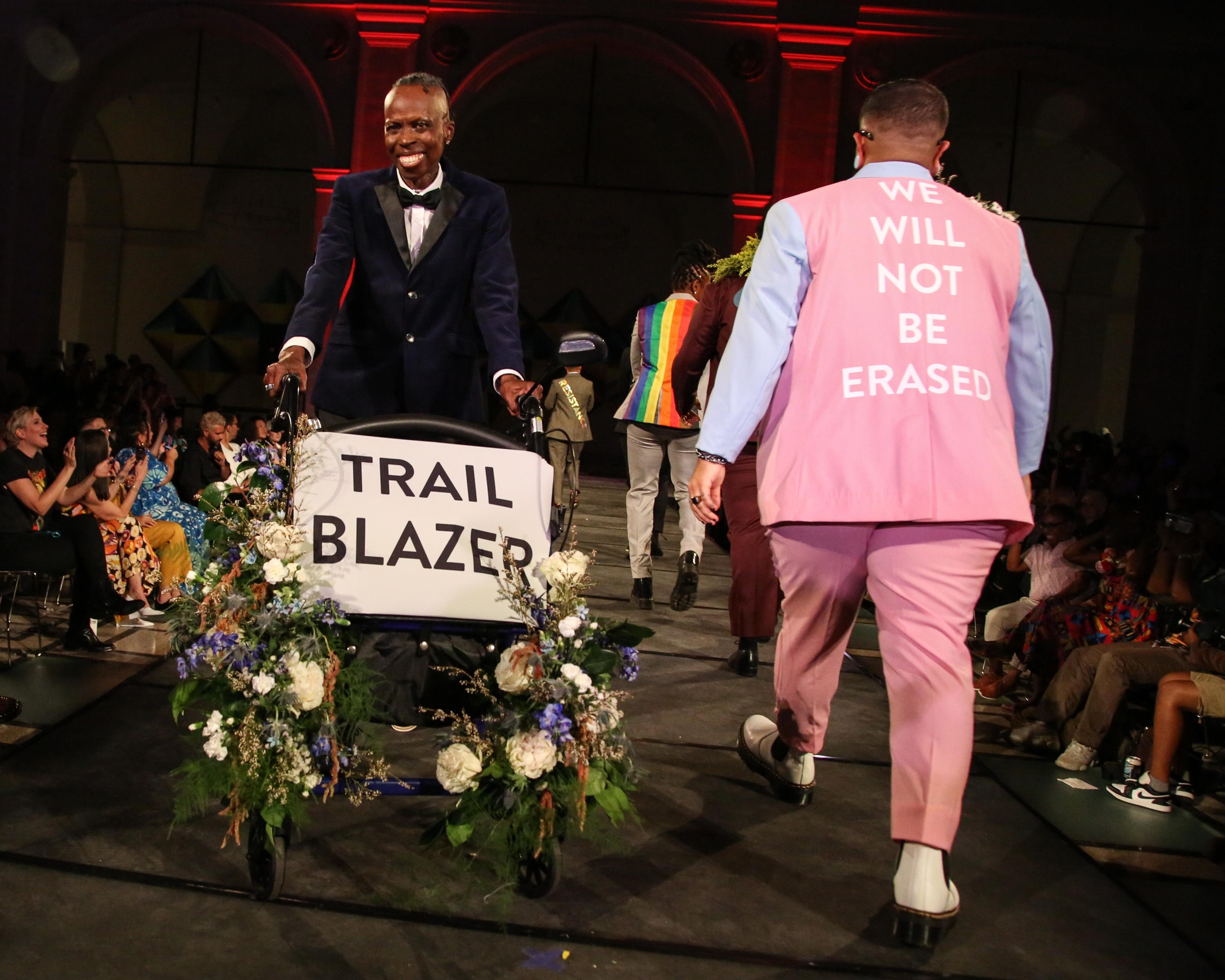 Models on a runway, one pushing a mobility device with flowers and a banner reading “Trail Blazer,” another wearing a jacket reading “We Will Not Be Erased”