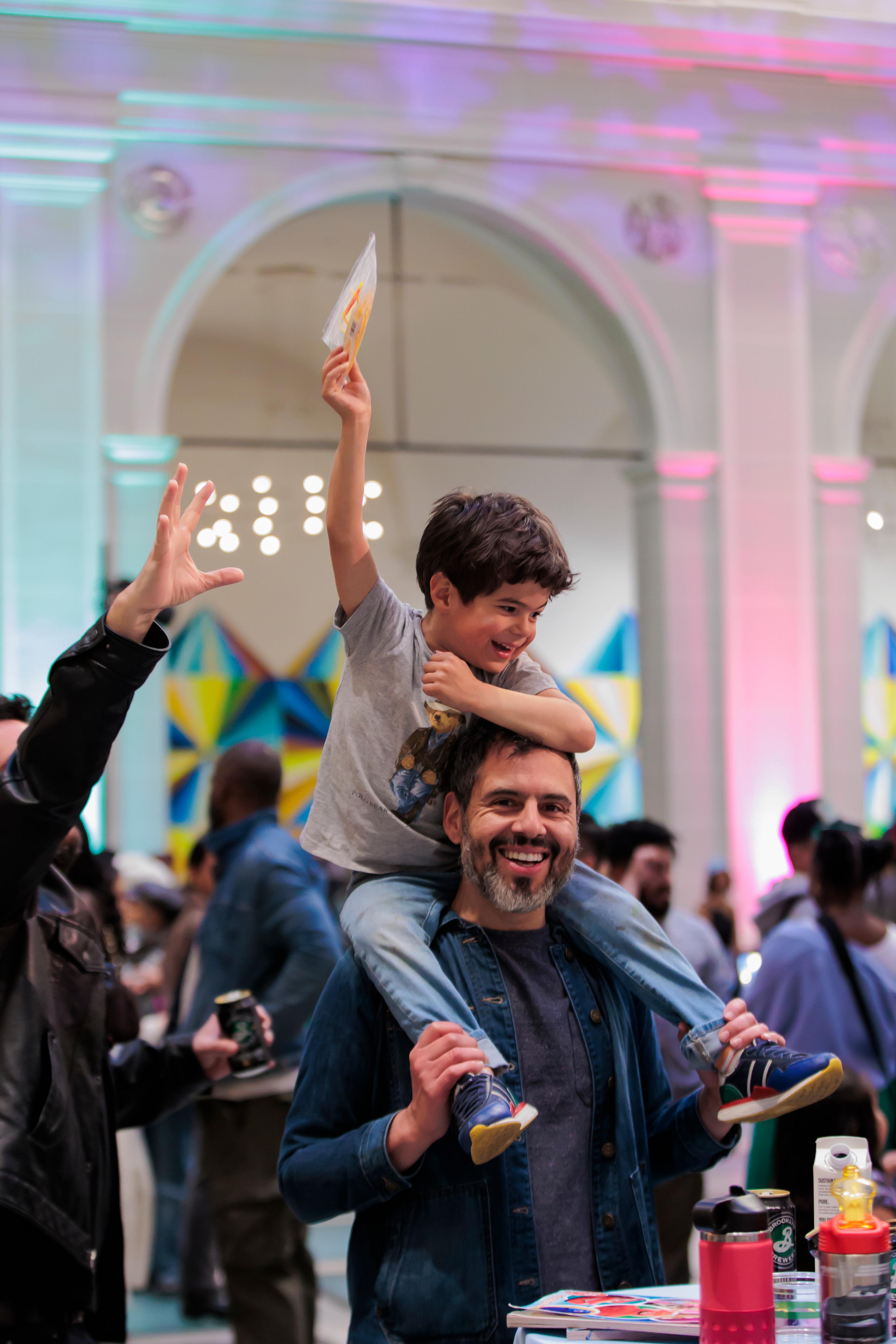 A man smiles and holds a child on his shoulders in a crowded art gallery