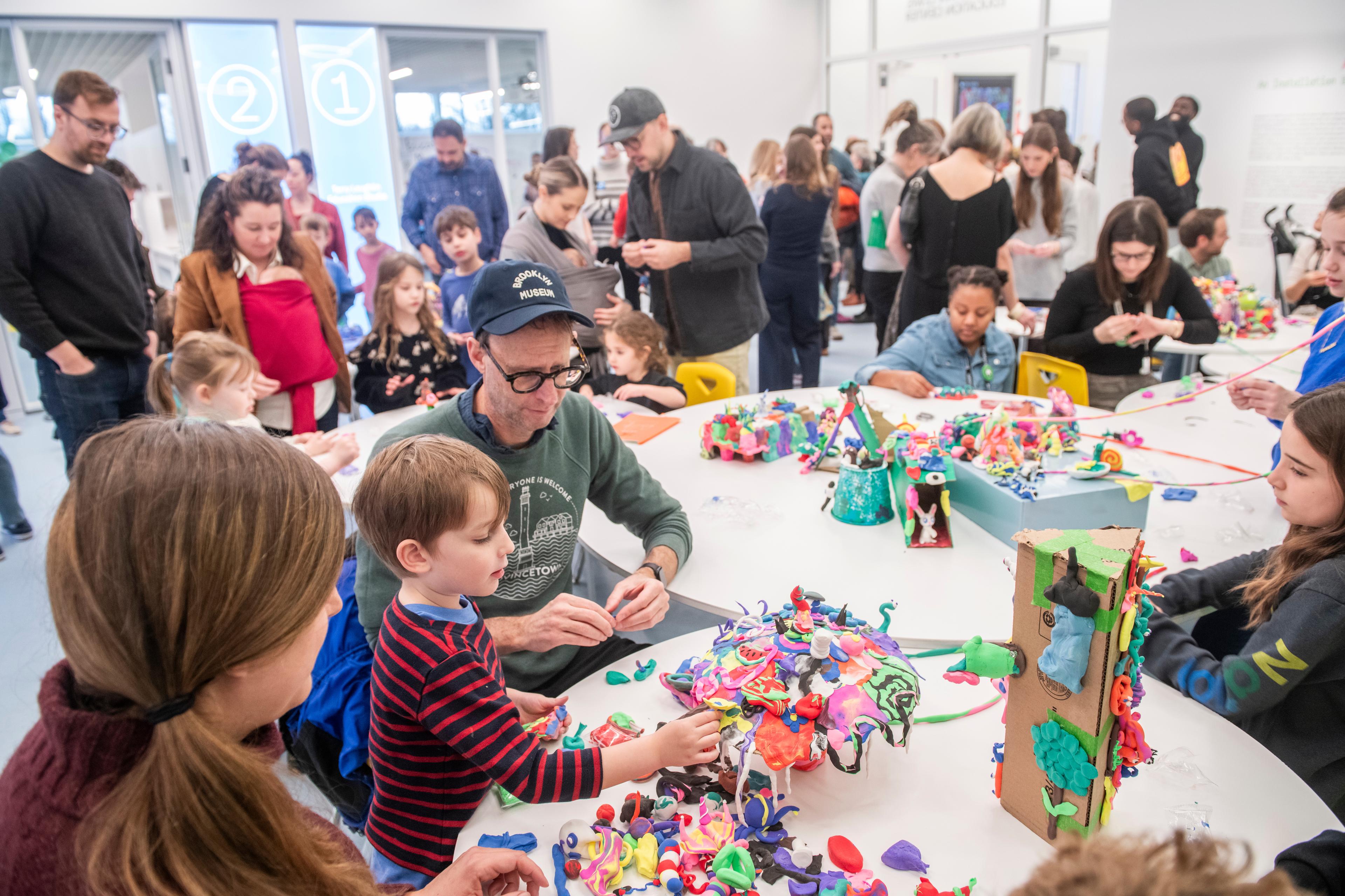 Multiple families sit around white round tables building sculptures out of play-dough