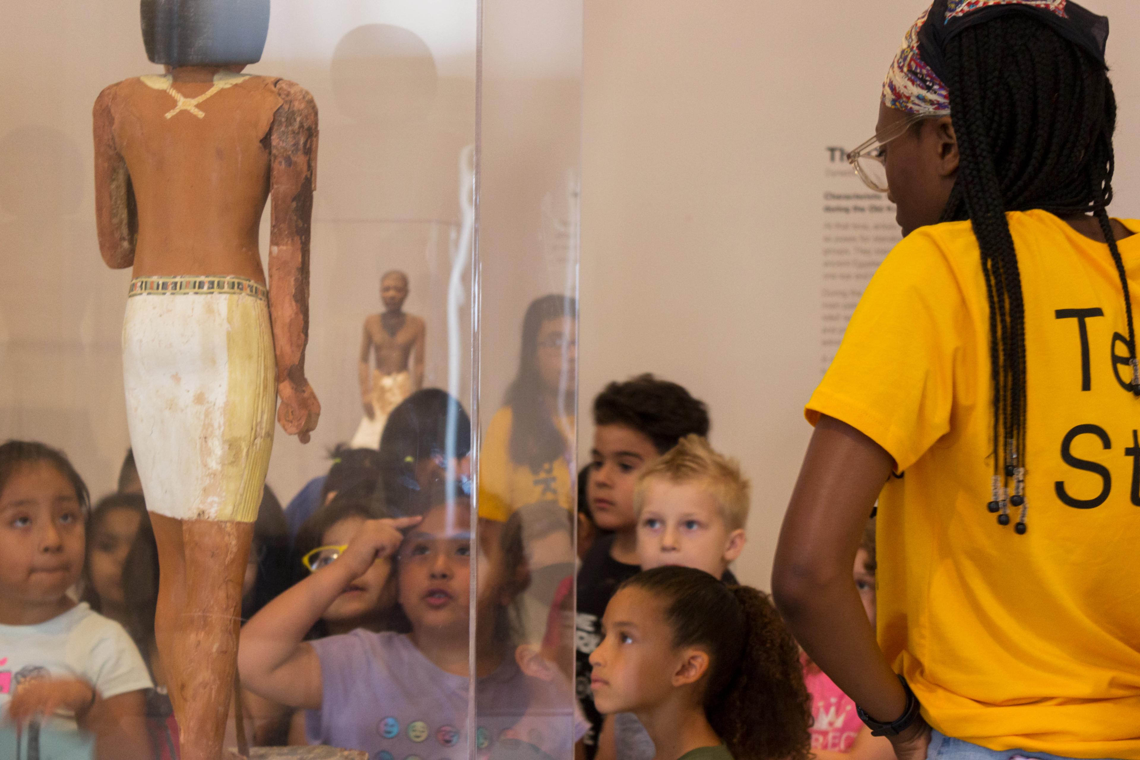 A group of young children surround an ancient Egyptian statue with a teen in "Teen Staff" shirt speaking to them