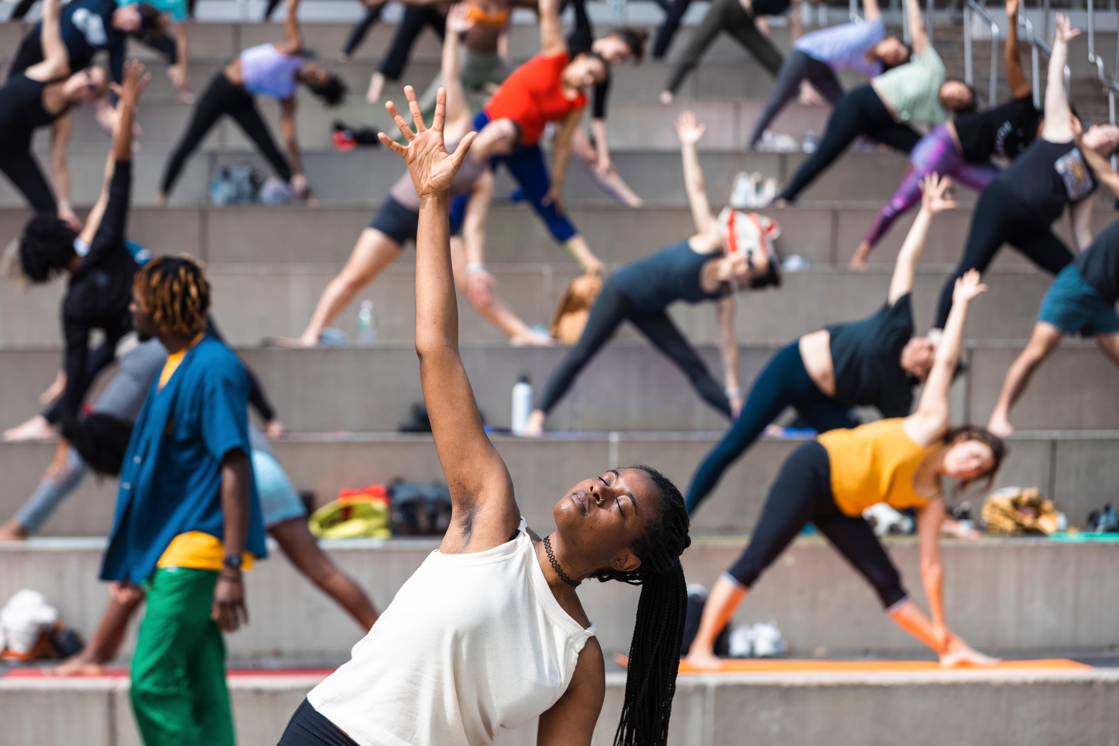 A yoga instructor poses with arm reaching up to the sky, with a dozen adult students replicating the pose behind them