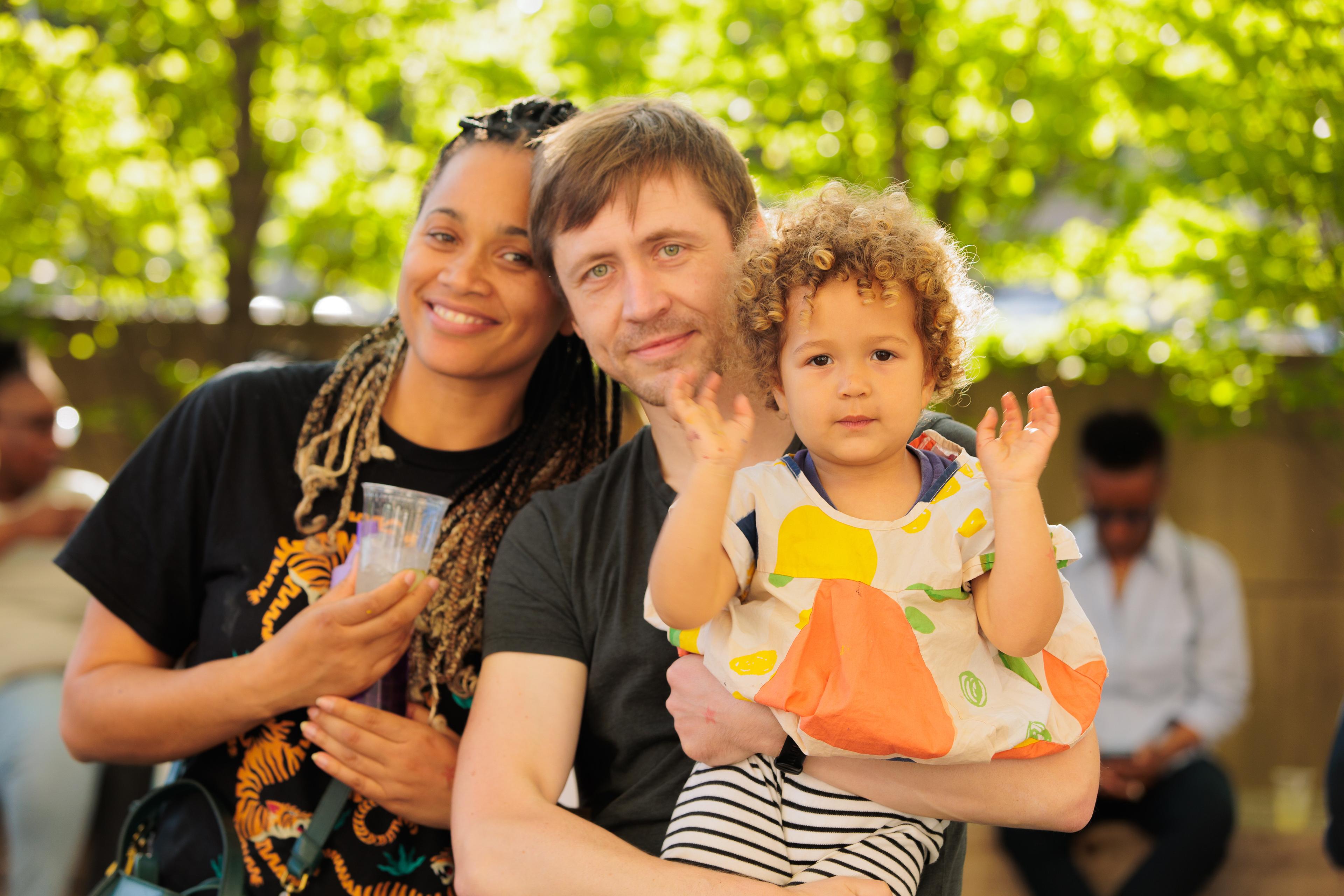 A family of three poses together, smiling outdoors. One parent is holding a young child in his arms.