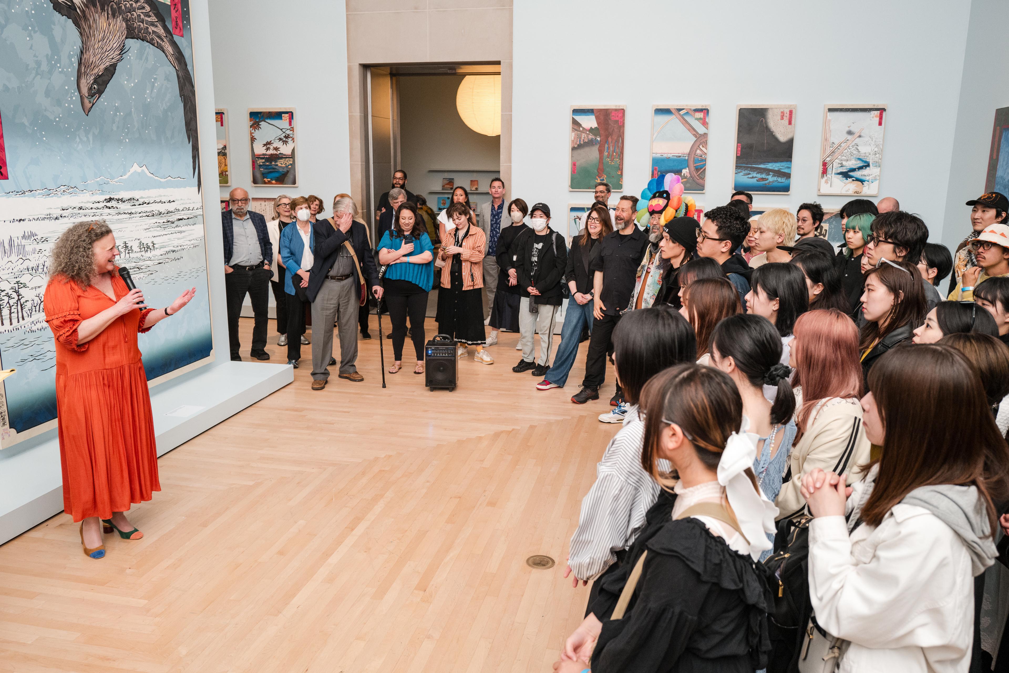 A person in an orange dress smiles and gestures while talking to a small audience in an art gallery