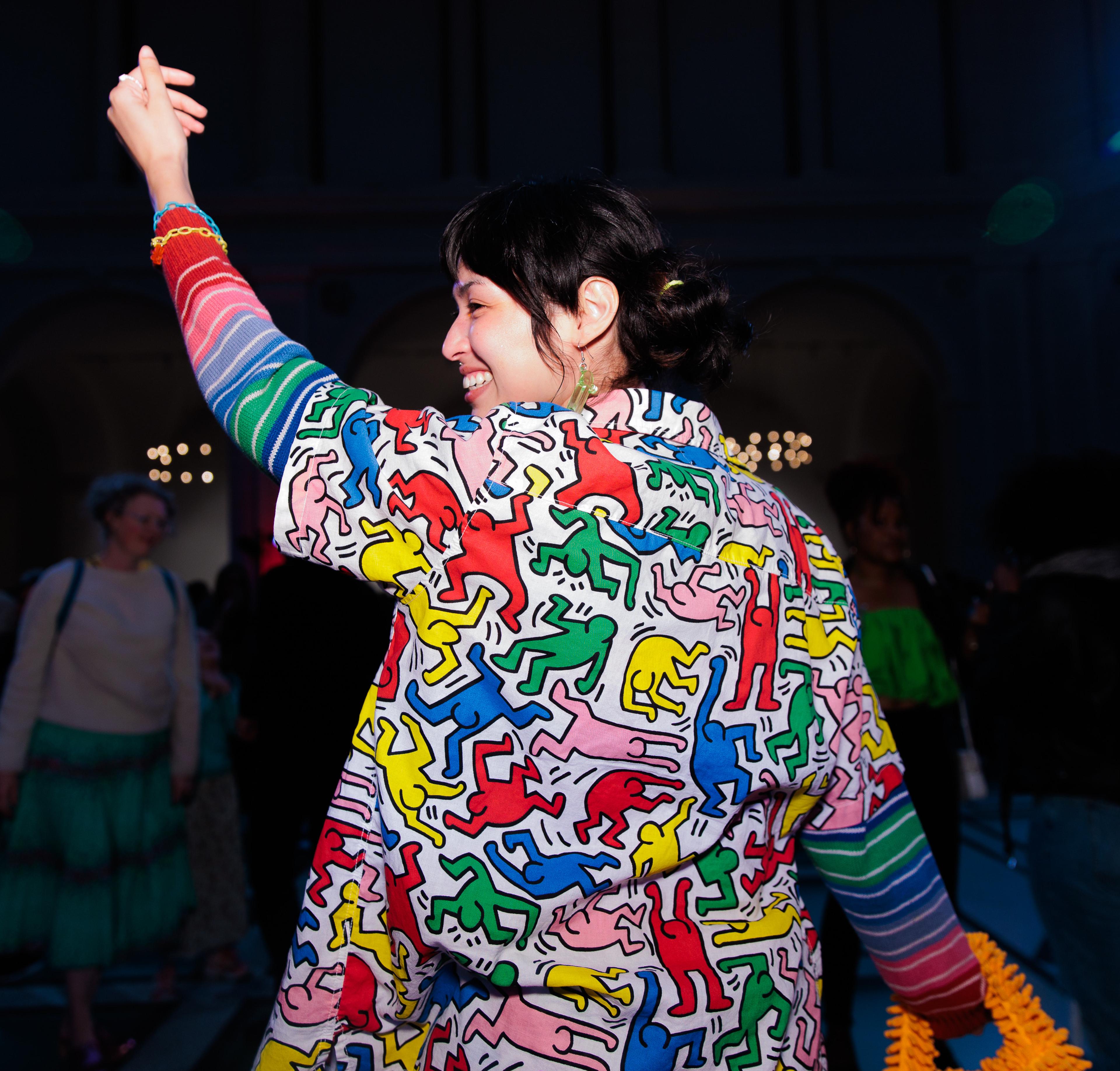 A young adult wearing a colorful shirt gesturing figures raises one hand up while dancing