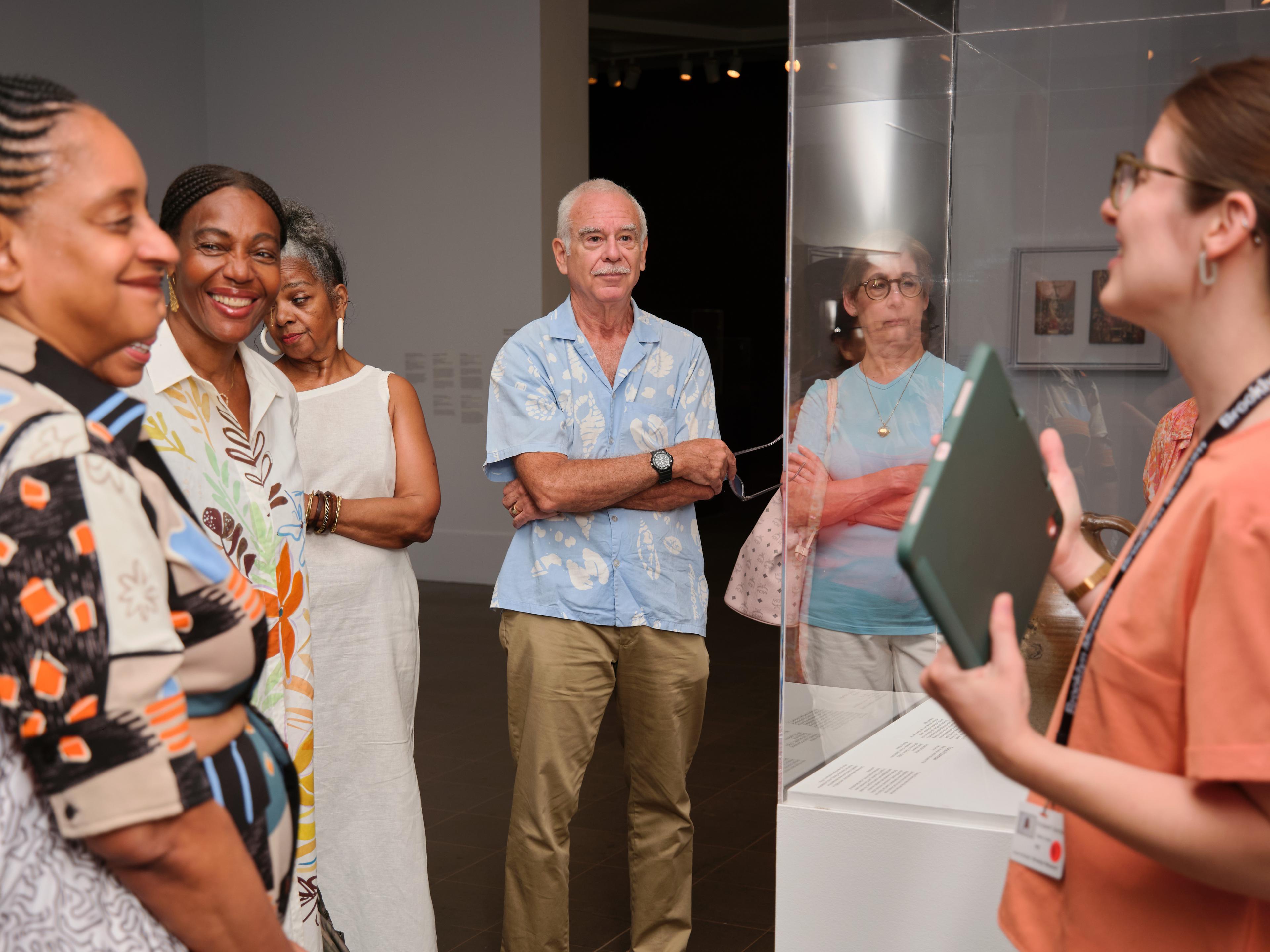 An adult holding an iPad speaks to a group of smiling adults around a case of artworks