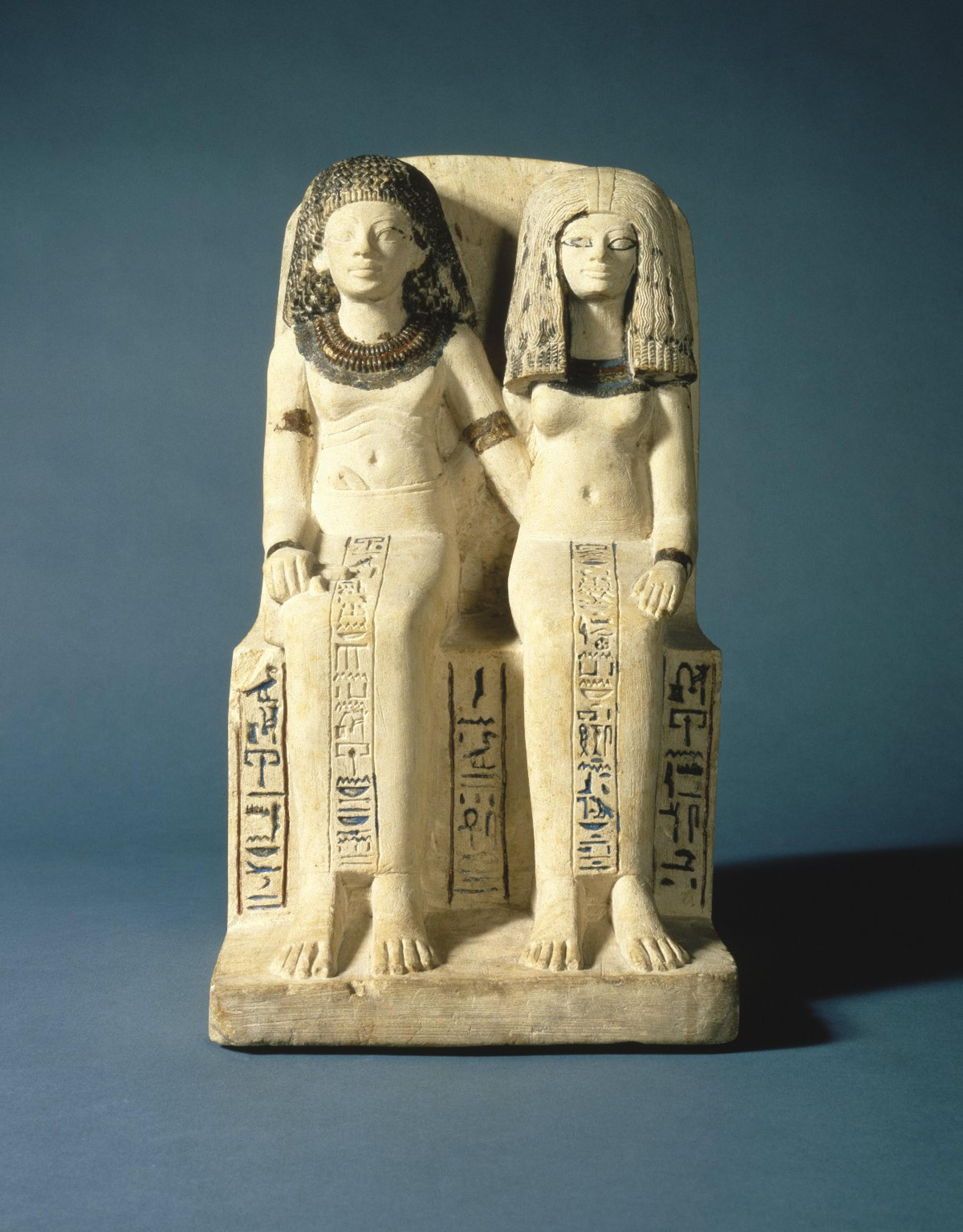 An ancient Egyptian sculpture of a seated couple wearing voluminous hairstyles and tight clothes with hieroglyphic writing