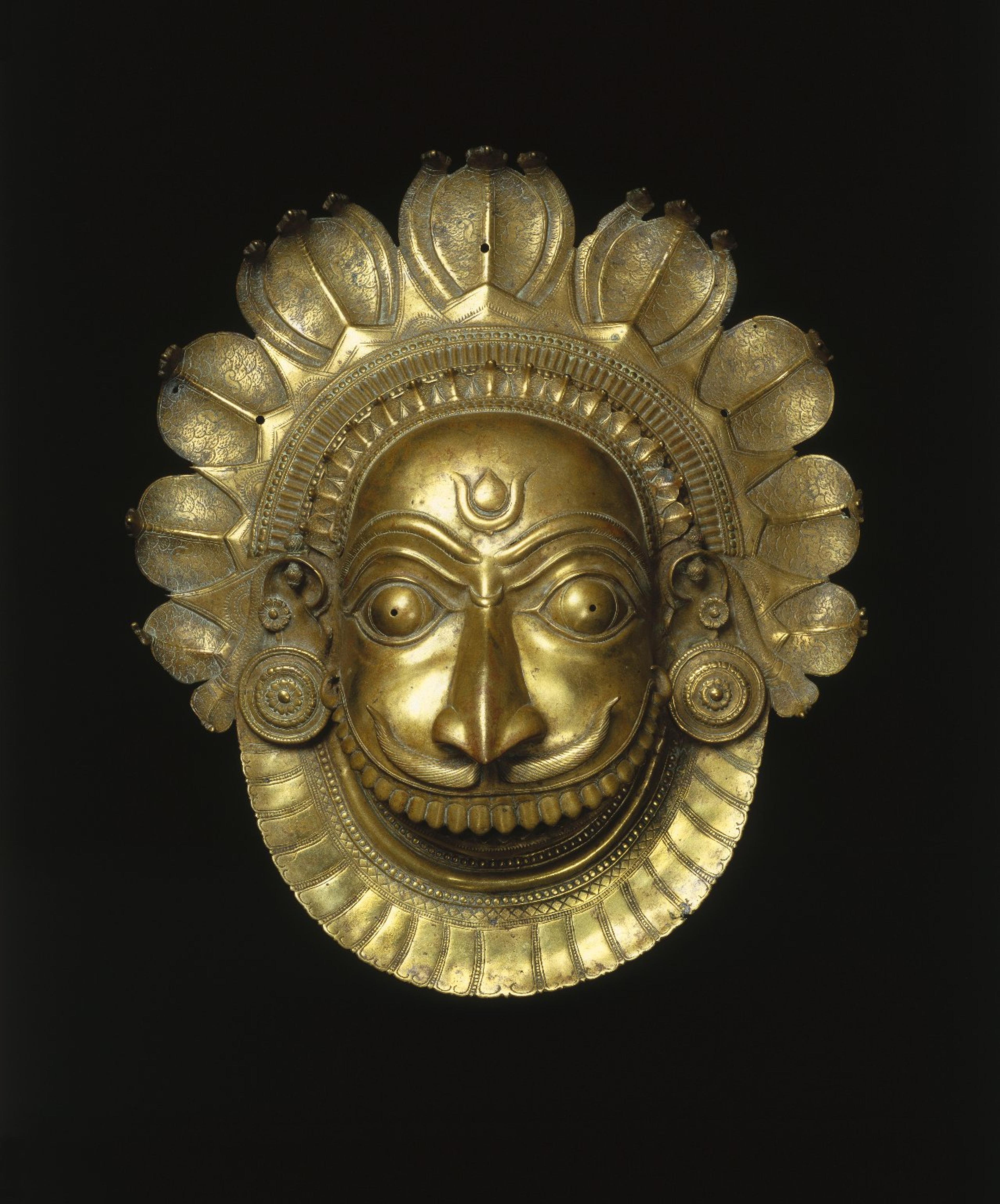 A brass mask features a humanoid face with wide eyes, a thin moustache, and an oversized mouth bearing numerous teeth; the head is crowned with dozens of cobras.