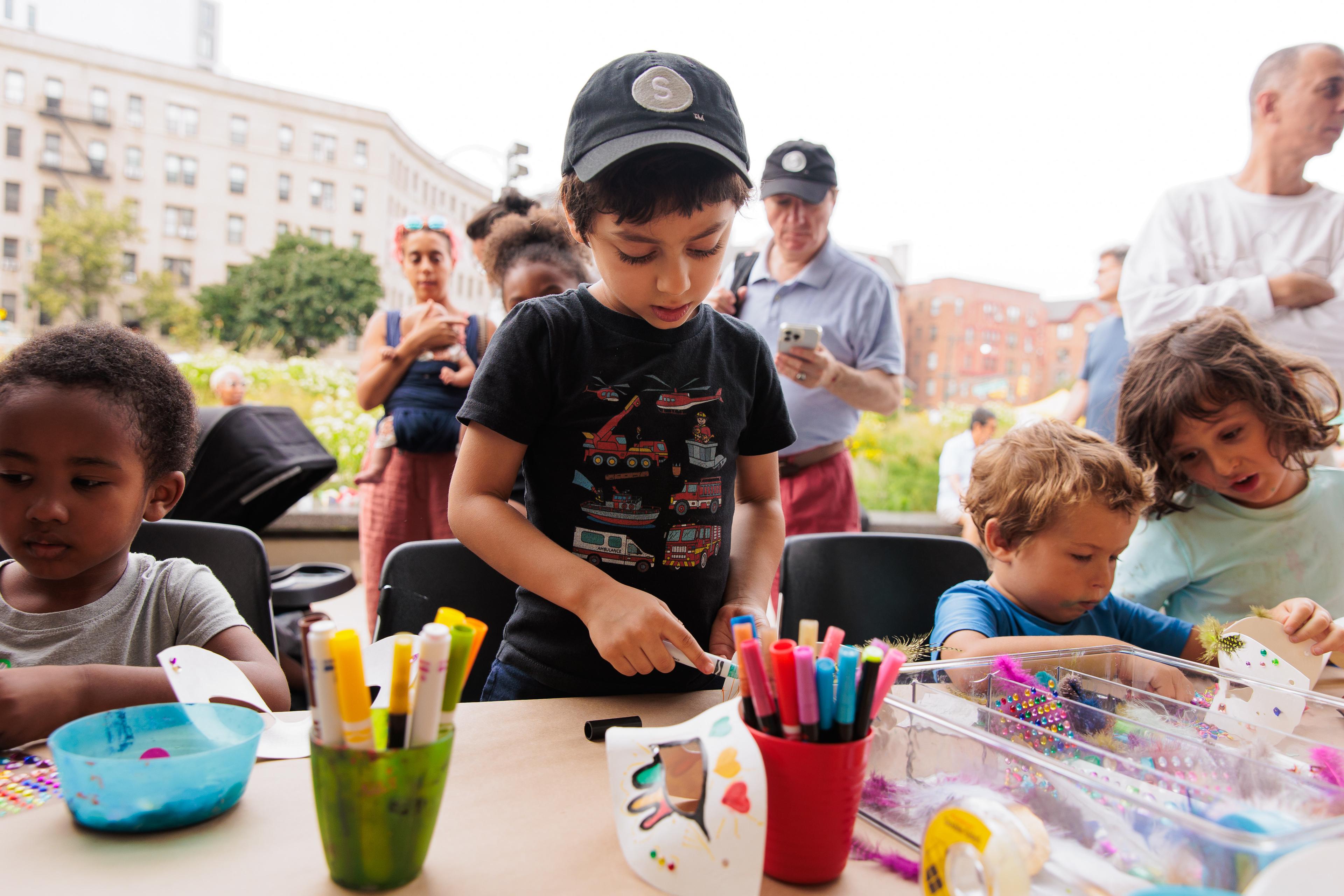 Children drawing with markers at a table outdoors, with adults standing behind