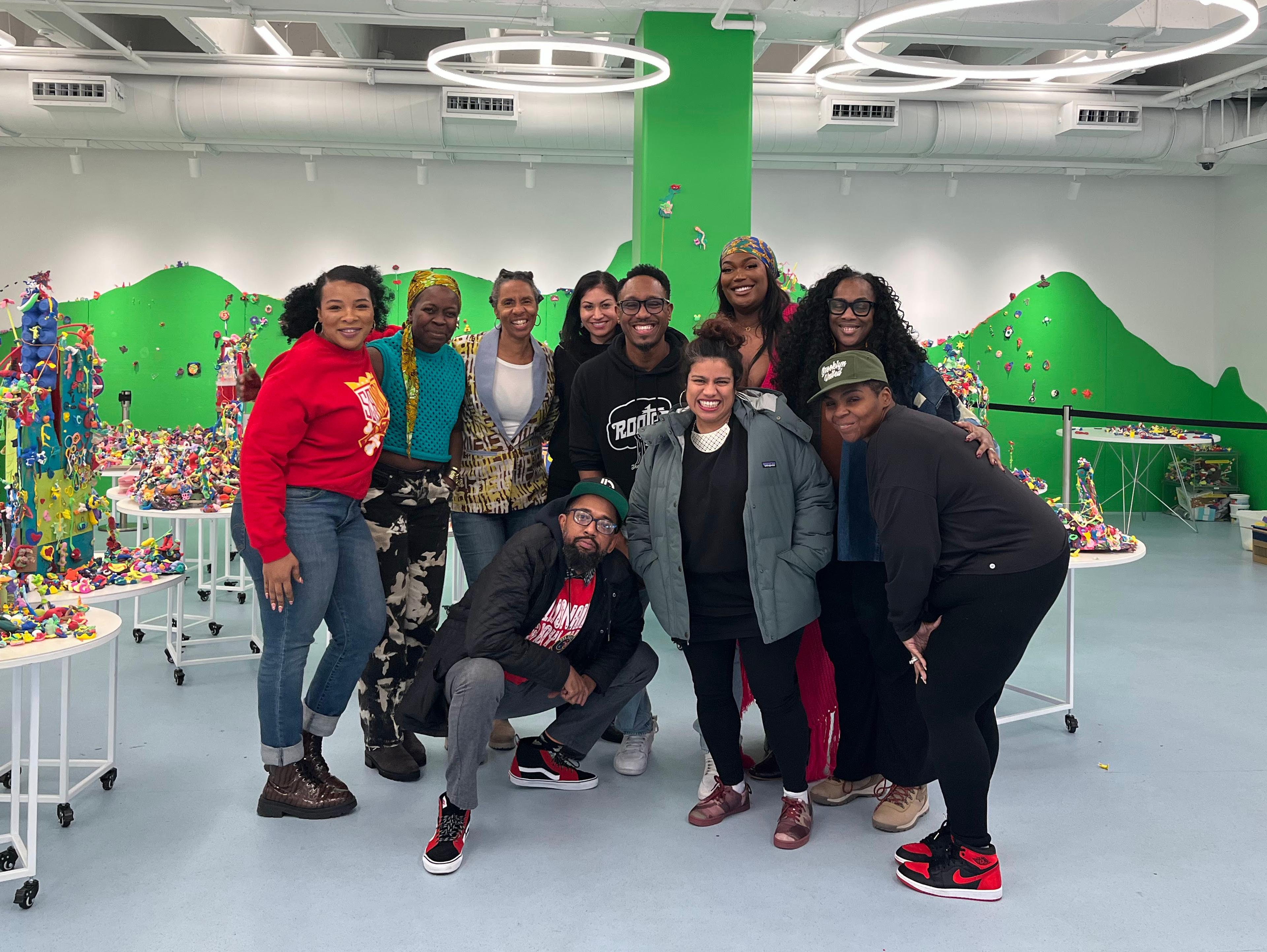 A group of ten adults pose together in a large bright room with green walls, and tables of playdough sculptures