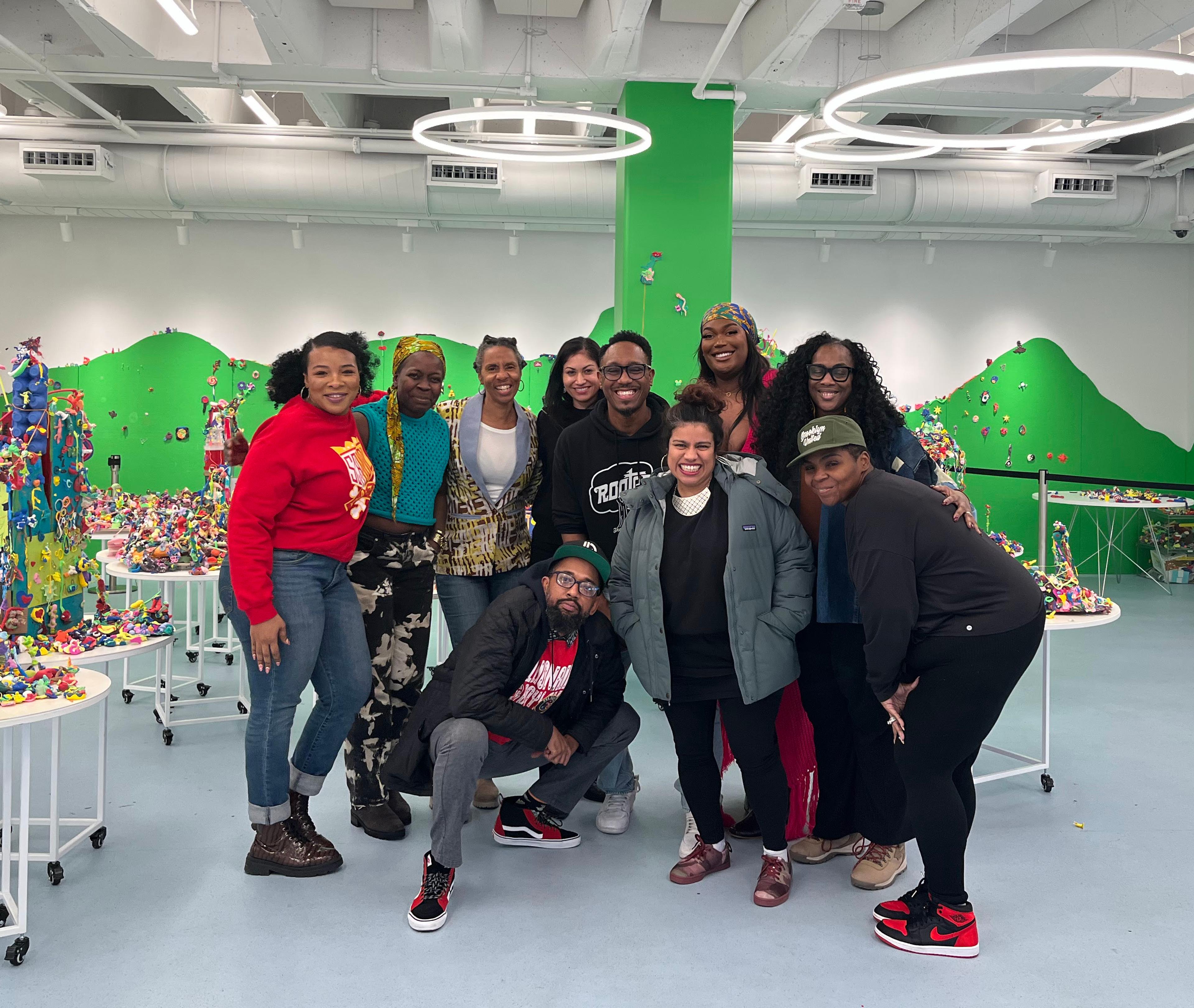 A group of ten adults pose together in a large bright room with green walls, and tables of playdough sculptures