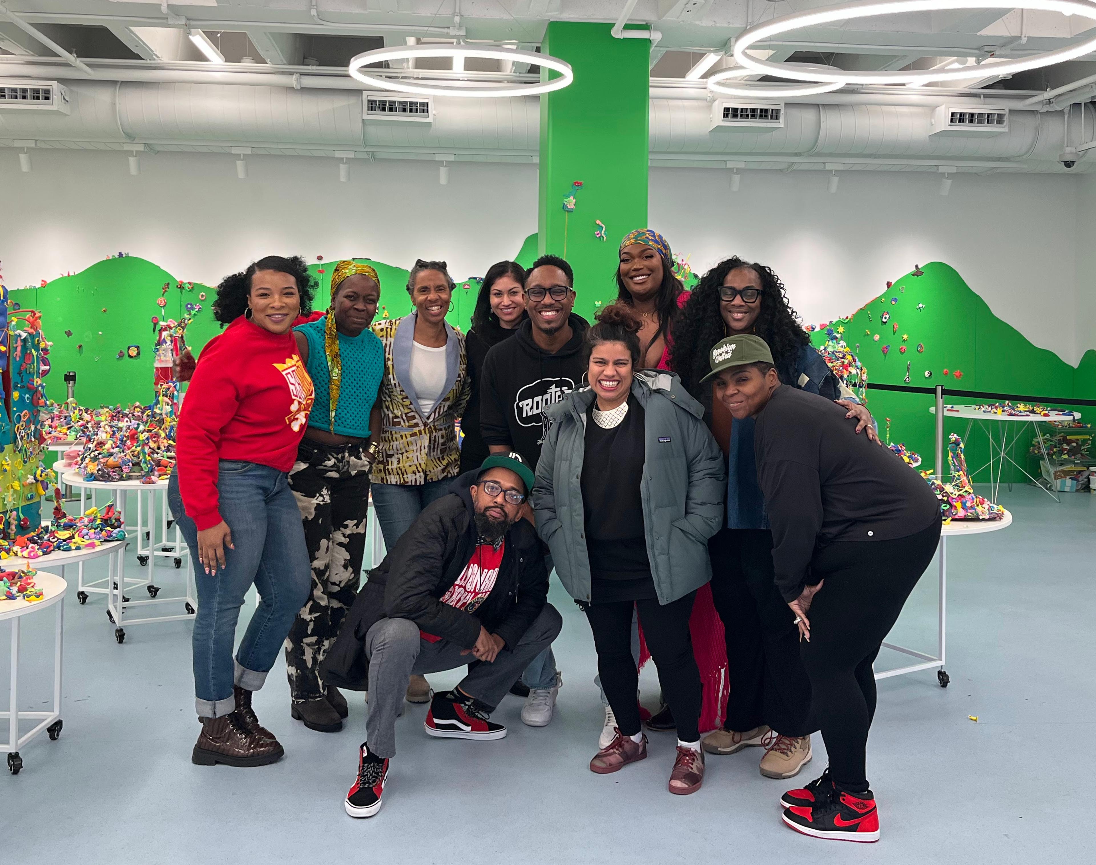 A group of ten adults pose together in a large bright room with green walls, and tables of playdough sculptures