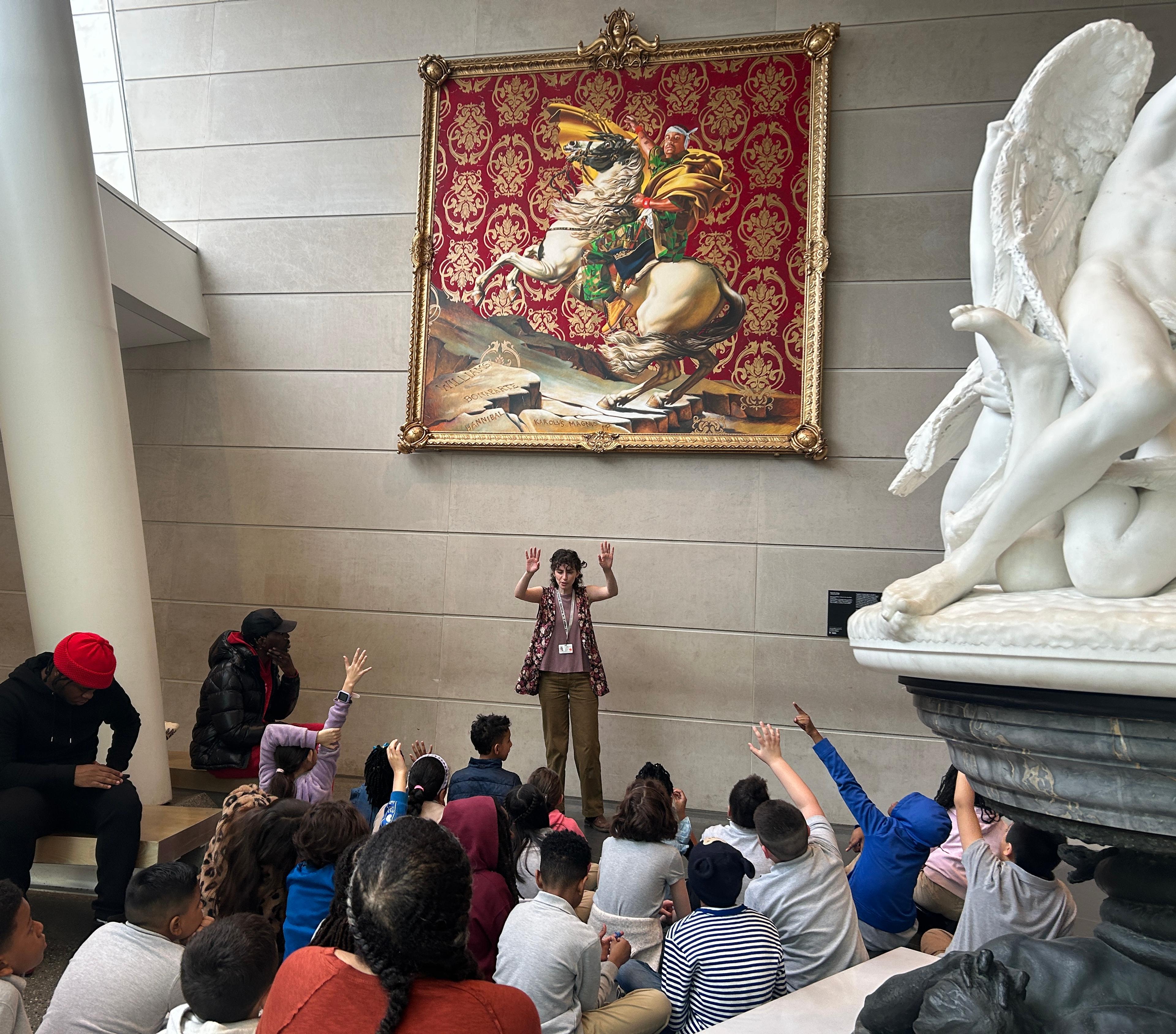 An adult instructor interacts with a seated group of young students, surrounded by a large painting and sculpture